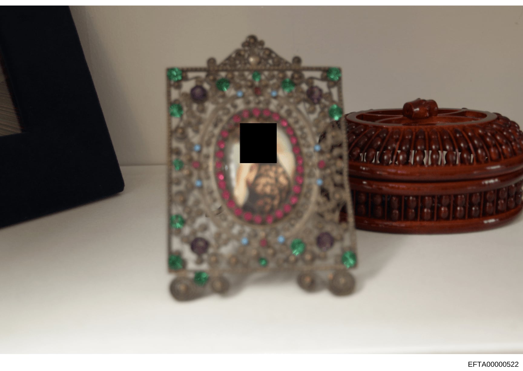 This photograph documents decorative items seized or photographed as evidence from an Epstein property. The image shows an ornate metal-framed object with gemstone inlays (featuring green, blue, and purple stones arranged around a central oval with a