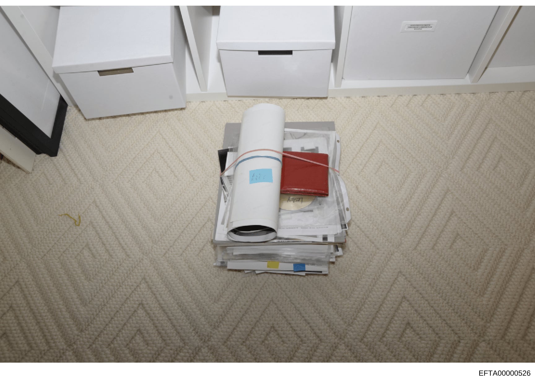 This is an overhead photograph of evidence collected during the investigation, showing a stack of documents, a rolled architectural or technical drawing with a blue label, a red leather-bound book or journal, and various papers arranged on a beige ca