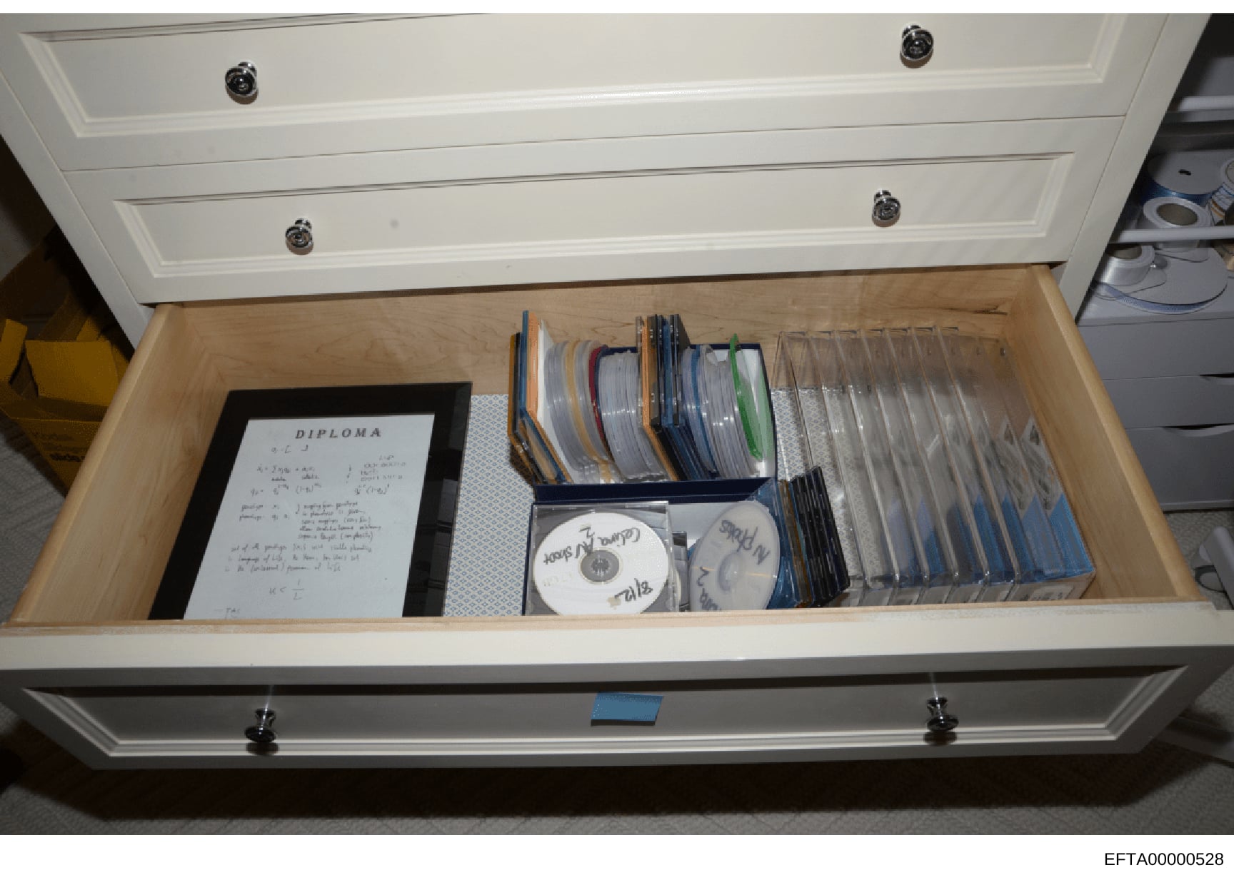 This photograph shows the contents of a white drawer seized during the investigation, containing a framed diploma document, numerous CDs organized in storage cases, and loose media. The items appear to be personal documents and digital materials stor