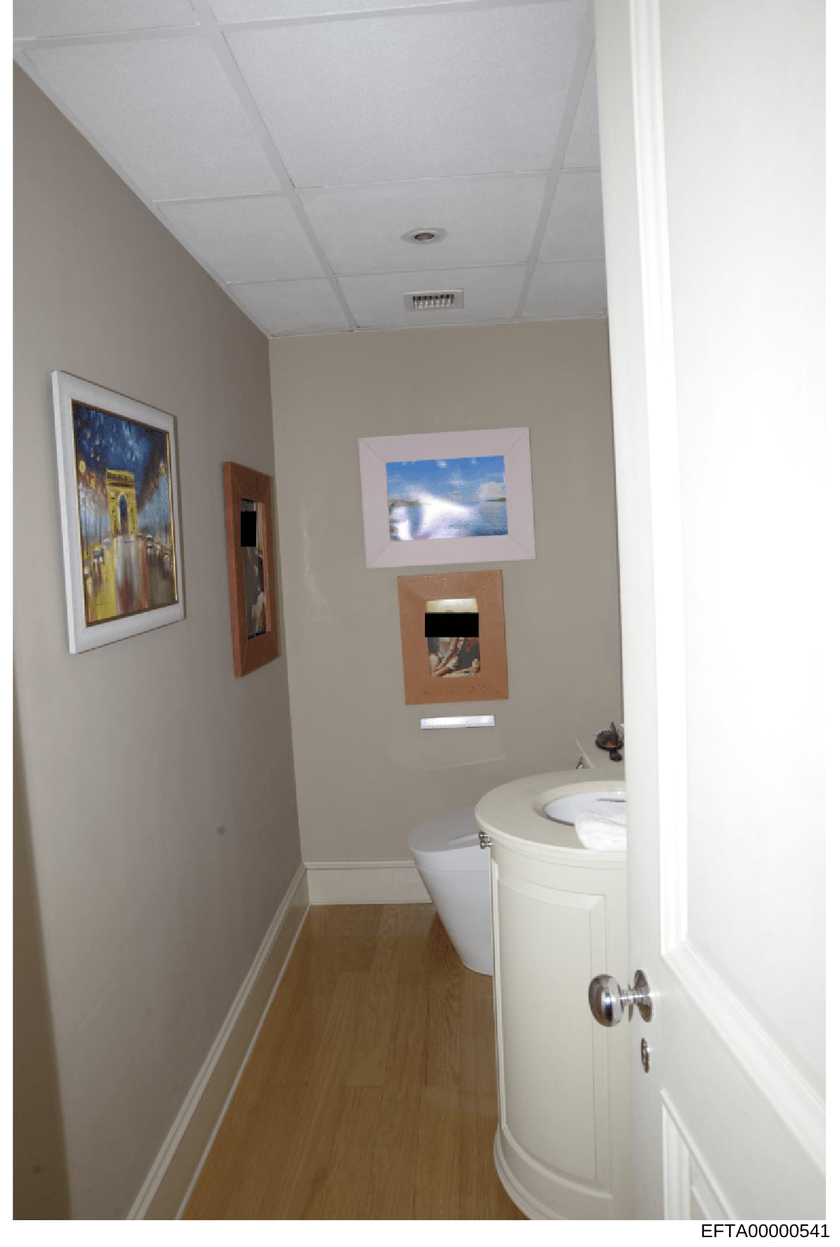 This is an evidence photograph depicting the interior of a bathroom, showing a toilet, sink vanity, and several framed artworks on the walls. The photo documents the interior condition and furnishings of a property relevant to the investigation. The