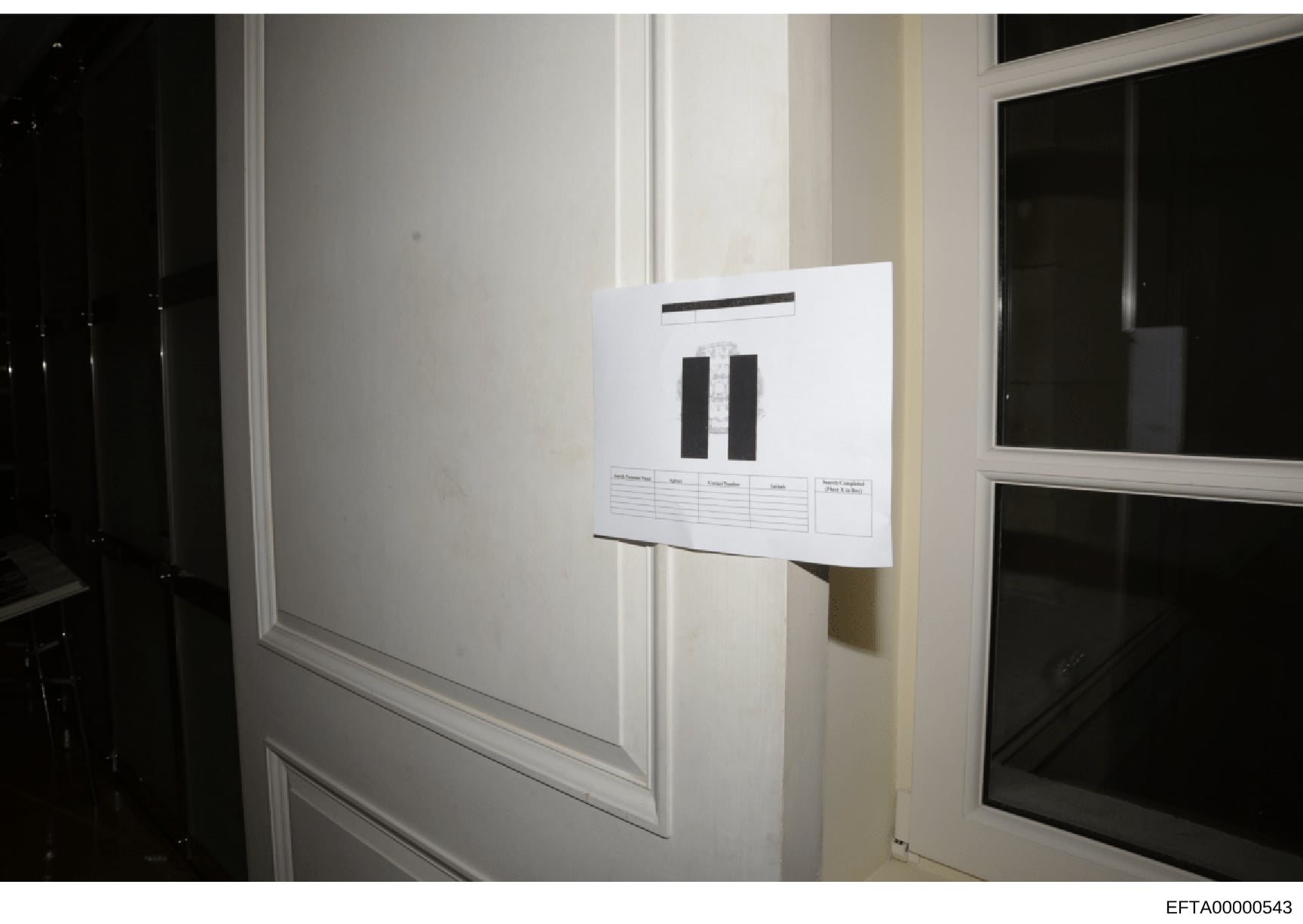 This is a photograph of an evidence item mounted on a white wall between two windows, displaying what appears to be two dark rectangular objects affixed to a white documentation board with a reference label and information grid. The photo was taken a