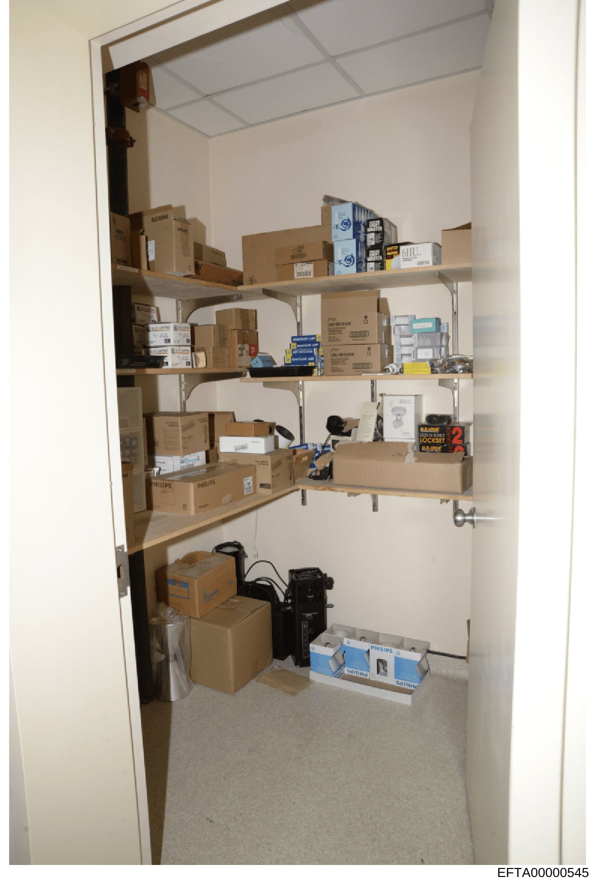 This is a photograph of a storage closet containing multiple cardboard boxes, electronic equipment, and miscellaneous items organized on metal shelving units. The image appears to be evidence documentation from the investigation, showing the contents