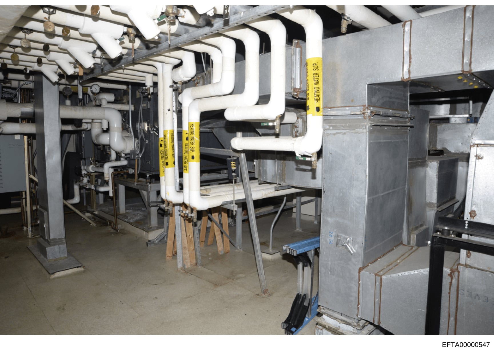 This photograph shows an interior view of a basement or mechanical room containing heating, ventilation, and air conditioning (HVAC) equipment along with extensive white PVC piping systems. The image appears to be documentation of building infrastruc