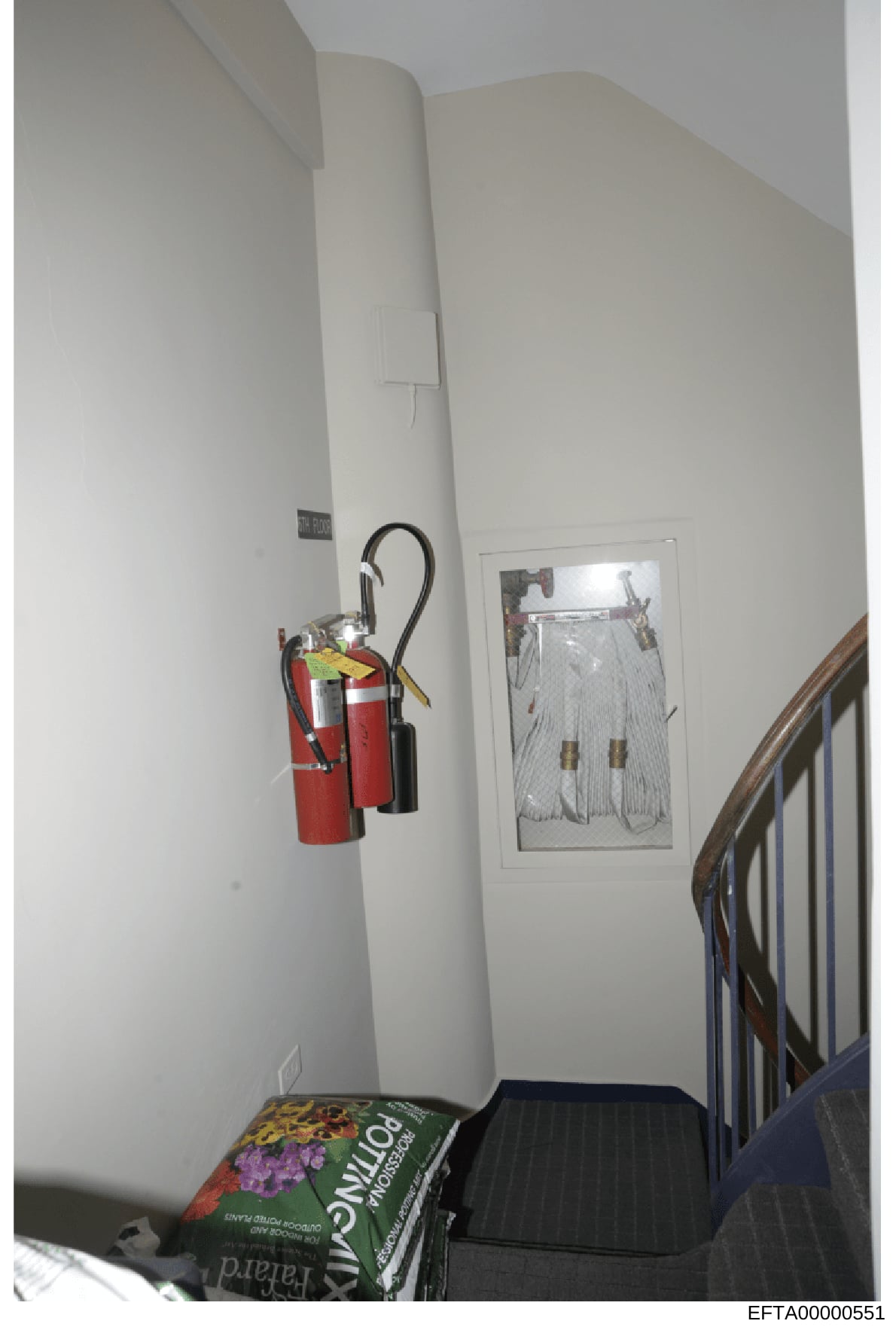 This is an evidence photograph showing an interior stairwell area, likely from one of Epstein's properties. The image captures fire safety equipment (two red fire extinguishers), electrical conduit, framed artwork on the wall, a wooden stair rail, an