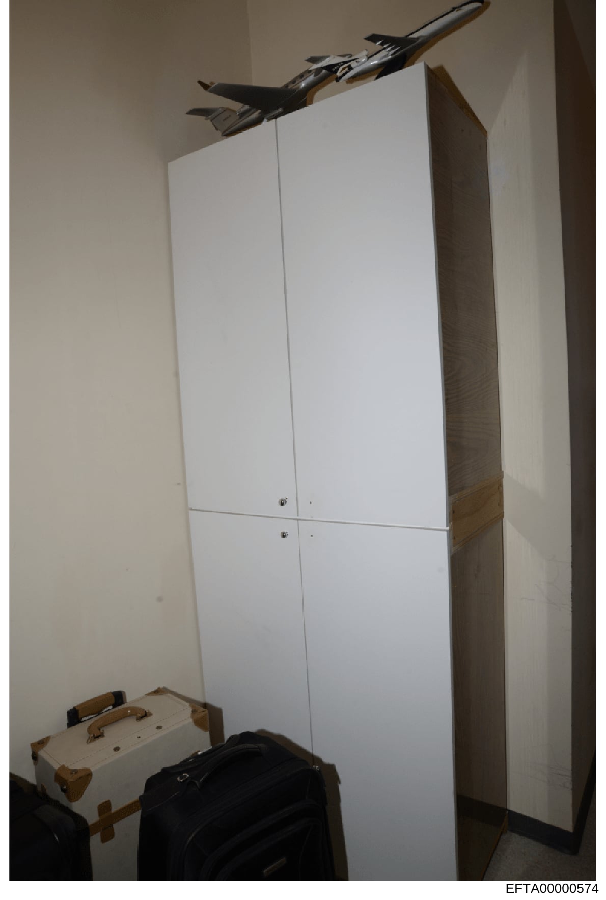 This is a photograph of an interior room or closet space showing a large white wardrobe/cabinet, beige walls, and luggage items on the floor. A model aircraft is mounted on the ceiling above the wardrobe. The image appears to be evidence documentatio