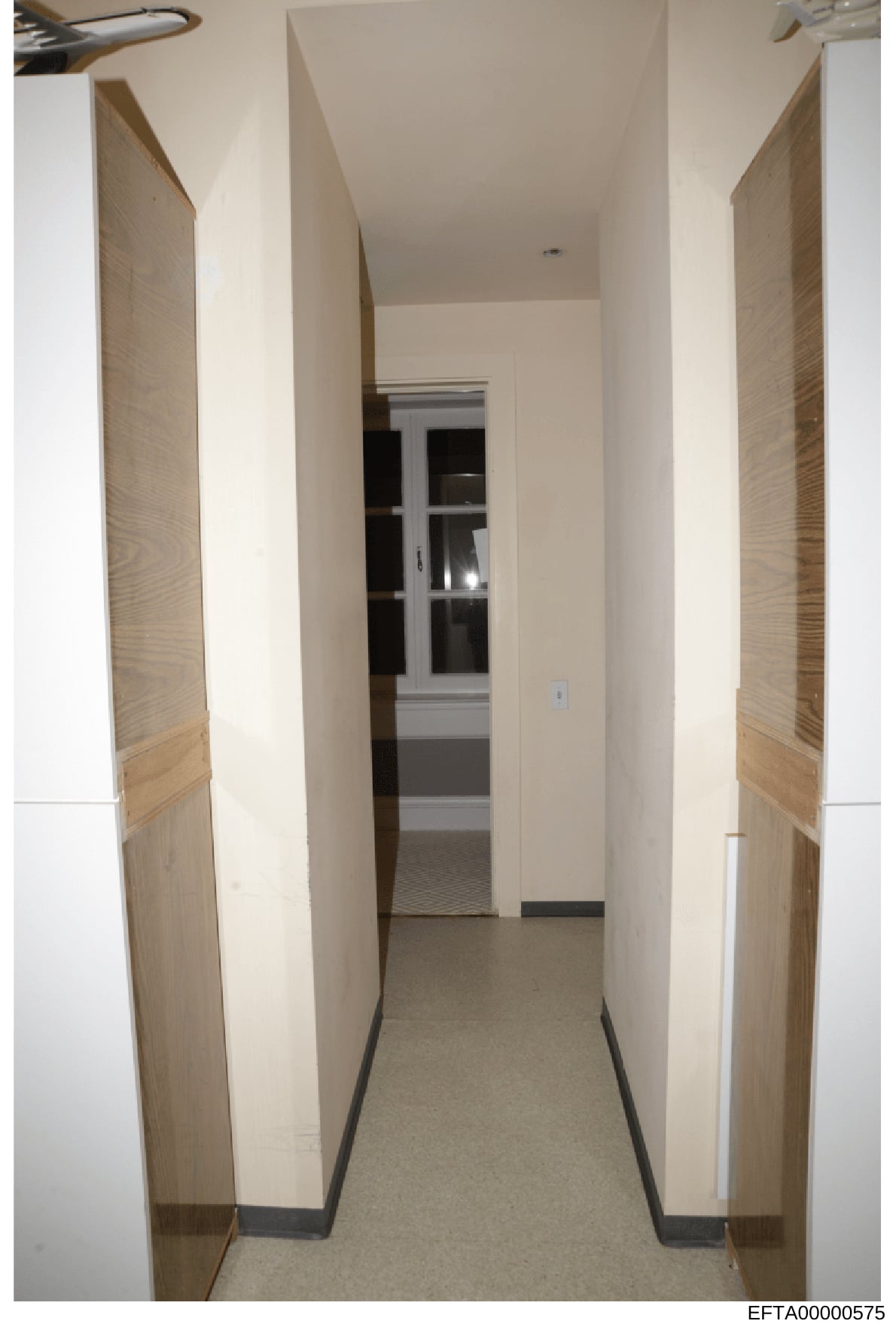 This is a photograph documenting the interior of a hallway within a property, showing clean, modern finishes with cream-colored walls, light wood doors on either side, and what appears to be a glass-paneled door or window at the end of the corridor. 