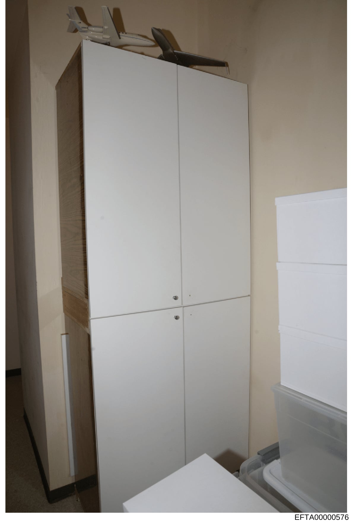 This is an evidence photograph showing a white four-door cabinet or wardrobe unit installed in what appears to be a residential interior space. The cabinet features white laminate doors with metal handles and appears to be positioned against beige/cr
