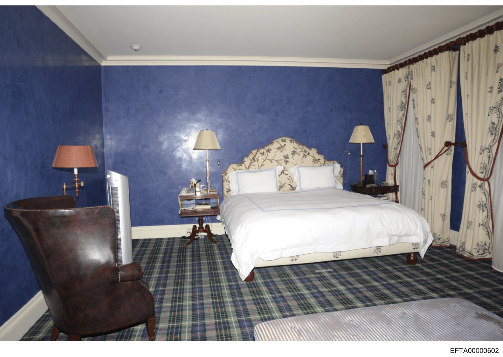 This is a photograph of an upscale bedroom interior, featuring blue walls, a cream-colored patterned headboard bed, formal furnishings including a leather wingback chair, nightstands with lamps, floral curtains, and a plaid carpet. The image appears 