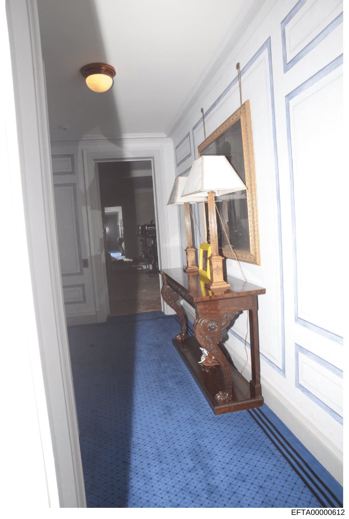 This photograph documents an interior hallway space, showing a decorated wooden console table with a framed mirror, table lamp, and decorative items. The hallway features white paneled walls, a blue patterned carpet, and recessed ceiling lighting. Th