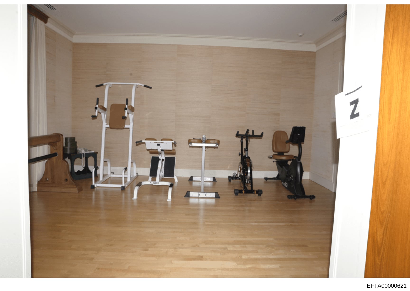 This photograph documents an interior home gymnasium containing various exercise equipment including stationary bikes, weight machines, and fitness apparatus. The room features hardwood flooring, neutral beige walls with crown molding, and was photog