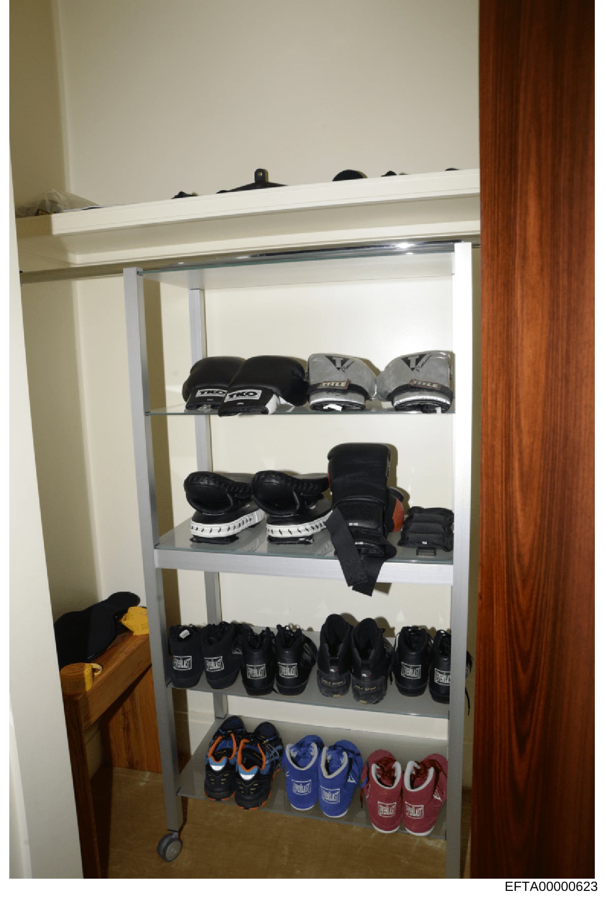 This is a photograph of a metal storage shelving unit containing approximately 20+ pairs of boxing gloves in various colors and sizes, along with other protective gear. The items appear to be organized on five shelves within what looks like a residen