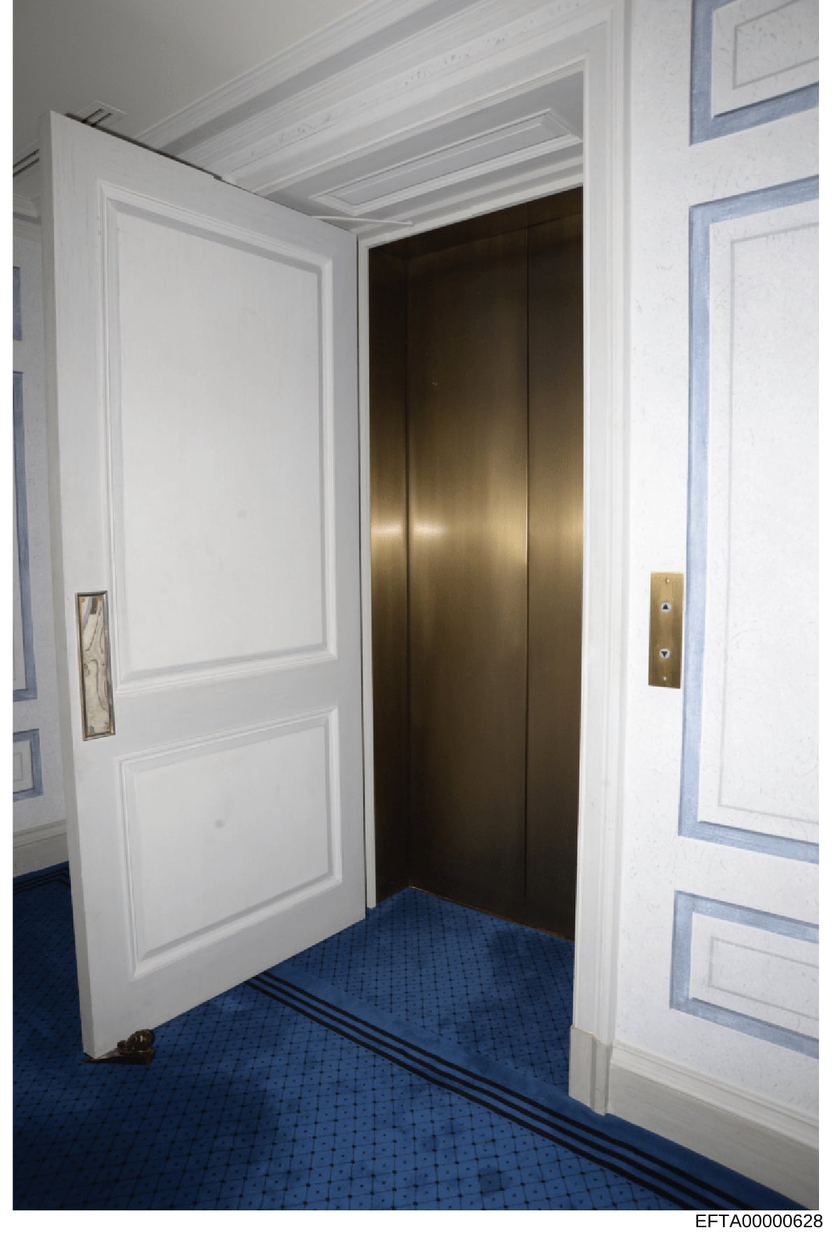 This is a photograph of an interior hallway, likely from one of Epstein's properties, showing an elevator entrance with dark bronze doors, white paneled walls, decorative ceiling detail, and blue carpeting. The image documents the interior design and