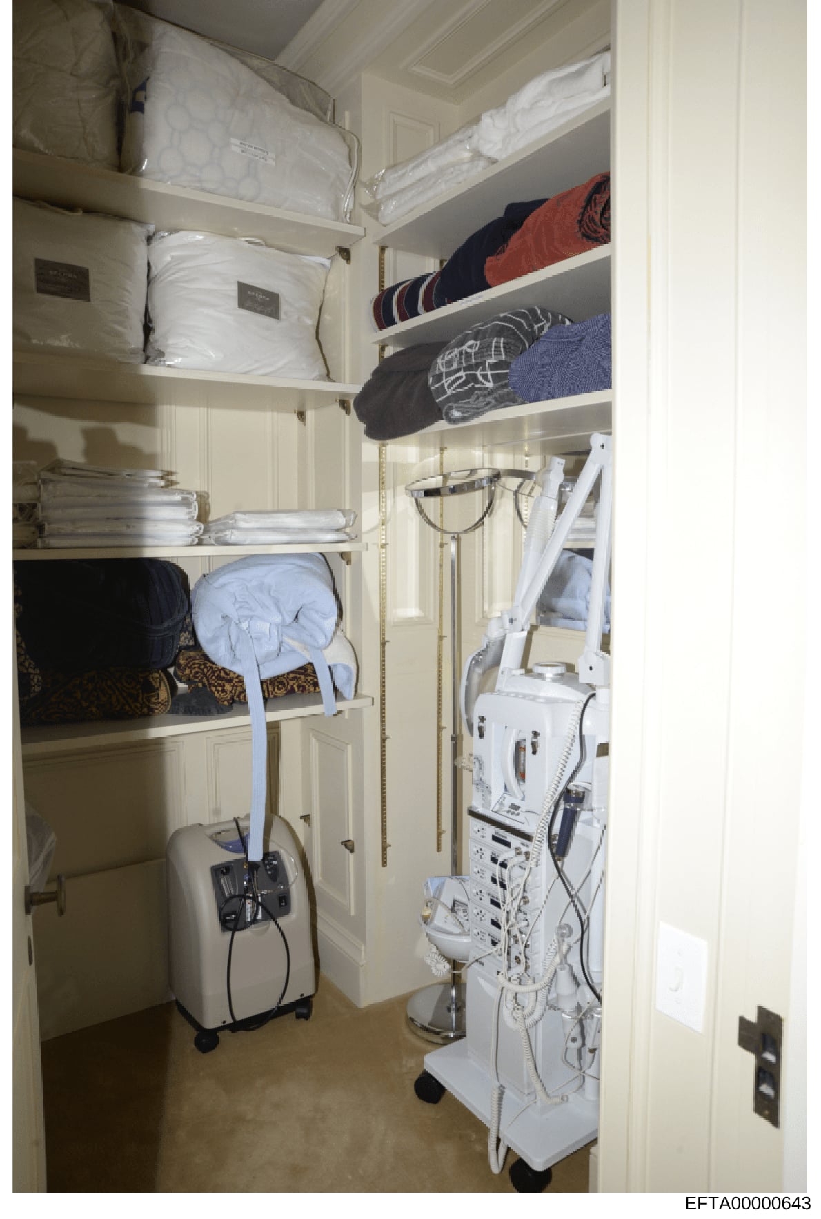 Evidence photograph showing the interior of a storage closet or linen room, containing shelves with bedding, linens, towels, and various household items. The photograph also documents white medical or electrical equipment stored on the right side of 