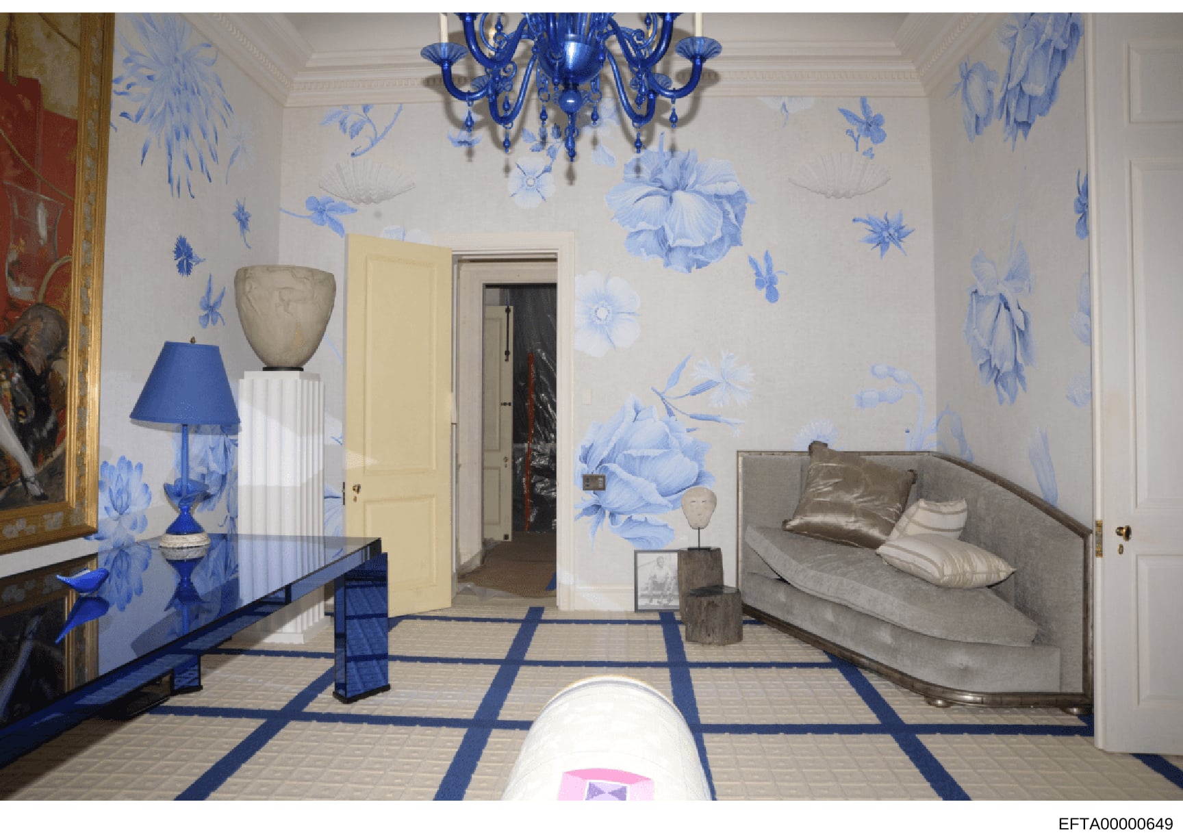 This is a photograph of an elegantly decorated bedroom featuring blue and white chinoiserie-style wall murals, a blue glass chandelier, and luxury furnishings. The room displays high-end interior design with botanical wall paintings, a neutral uphols