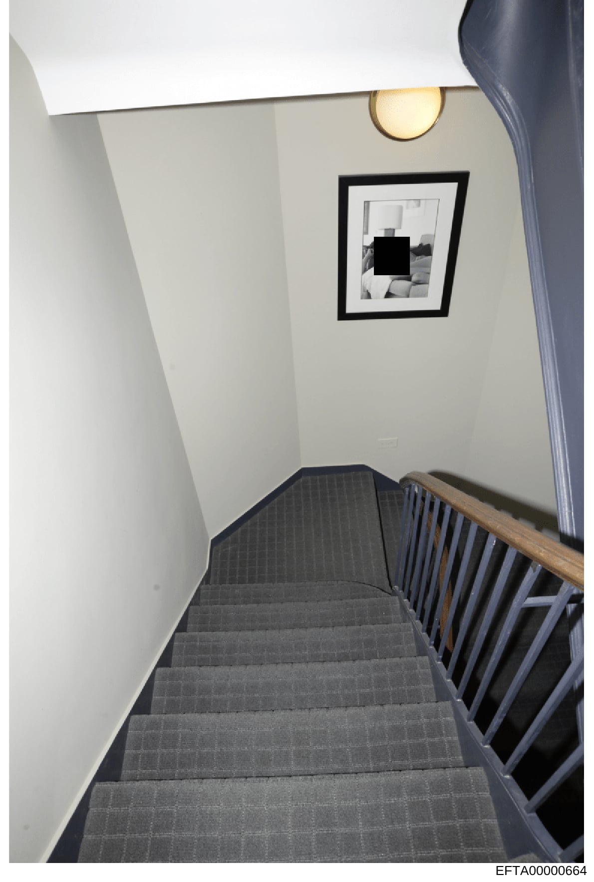 This is an evidence photograph showing an interior stairwell with clean, minimalist décor. The image depicts gray carpeted stairs with a wooden handrail, white walls, a round brass wall sconce, and a framed black-and-white photograph on the wall (par