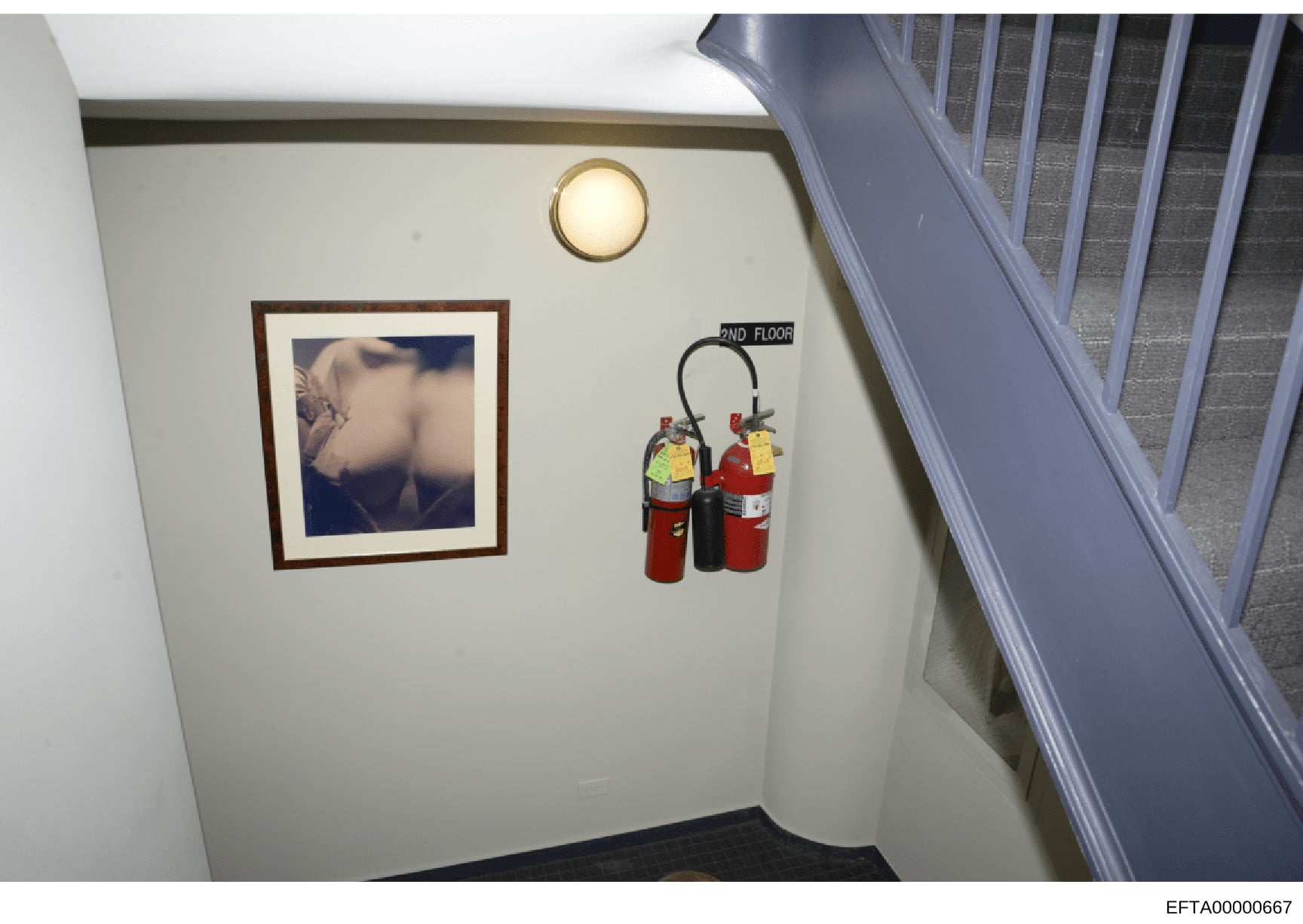 This is an evidence photograph showing the interior of a stairwell, likely from one of Jeffrey Epstein's properties. The image captures a cream-colored wall with a framed photograph, a circular ceiling light fixture, fire safety equipment, and a stai