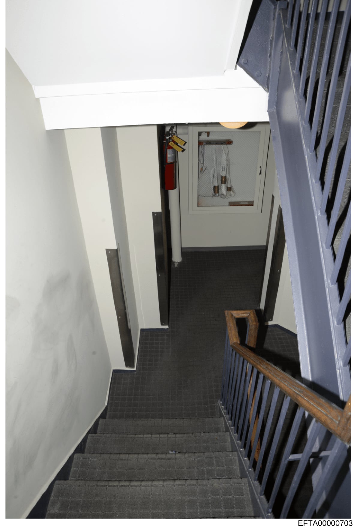 This is an evidence photograph showing a stairwell leading down to a basement or lower level area of a property. The image depicts white-painted walls, dark tiled flooring, a wooden railing, utility elements including a red fire extinguisher, and wha