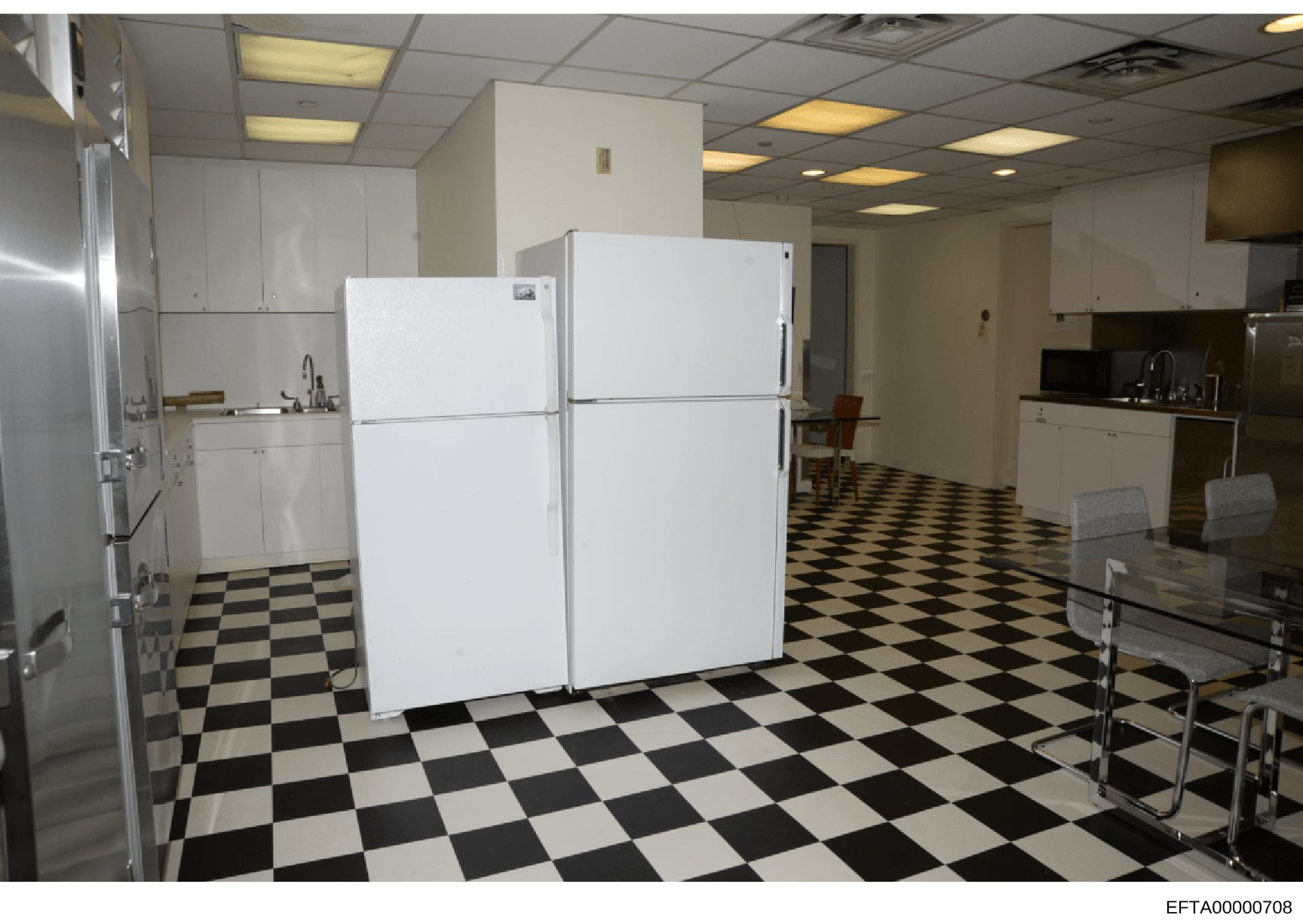 This photograph shows a commercial or institutional kitchen space, featuring white cabinetry, stainless steel appliances, and two large white refrigerators. The space has a distinctive black-and-white checkered floor and recessed ceiling lighting. Th