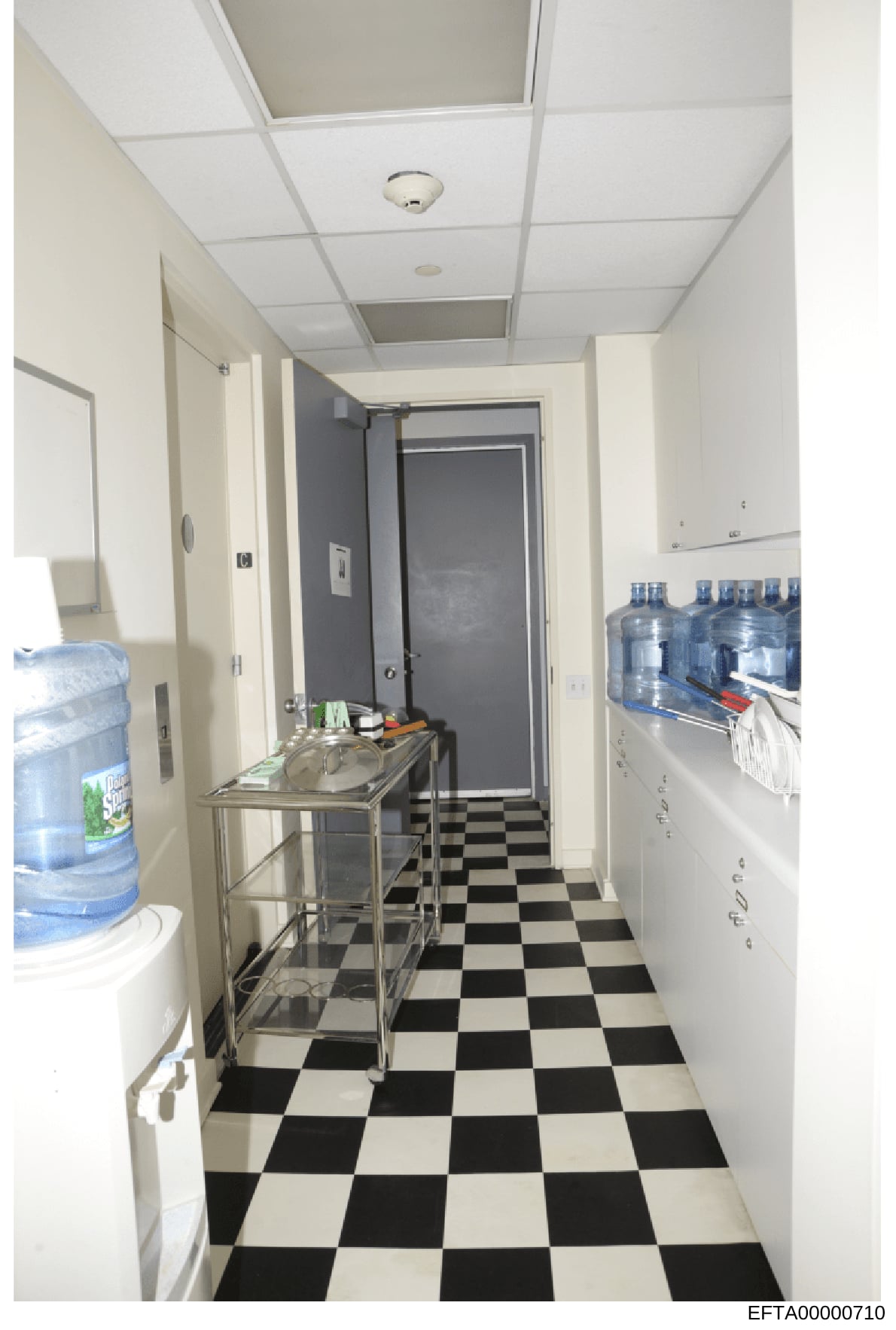 This photograph depicts an institutional-style hallway or break room with black and white checkered flooring, white drop-ceiling tiles, and commercial fixtures. The space contains water coolers, a stainless steel sink/counter unit, storage shelves wi