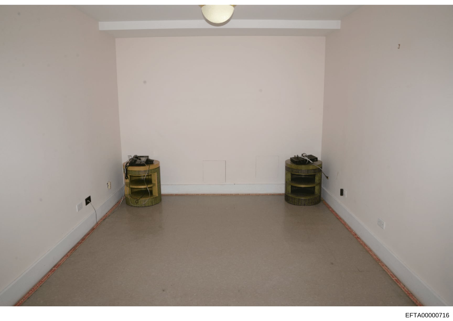 This is an evidence photograph showing an empty interior room with white walls and a concrete floor. Two cylindrical storage containers with olive/tan colored mesh material are positioned on opposite sides of the room. The room appears to be under re