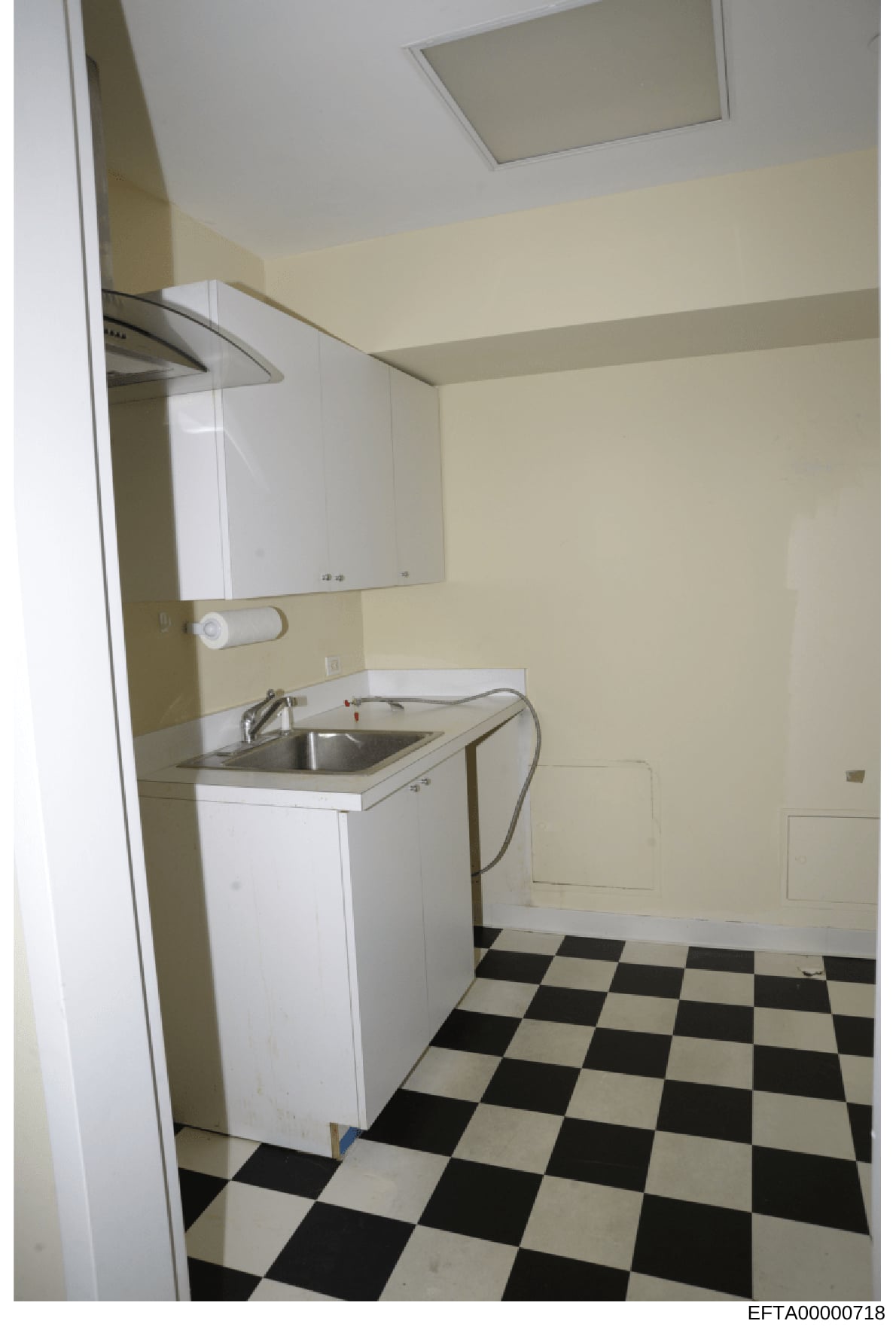 This is a photograph of a bathroom or utility room interior, showing basic fixtures and finishes. The image depicts cream-colored walls, white cabinetry, a stainless steel sink, and a distinctive black-and-white checkered tile floor. The photograph a