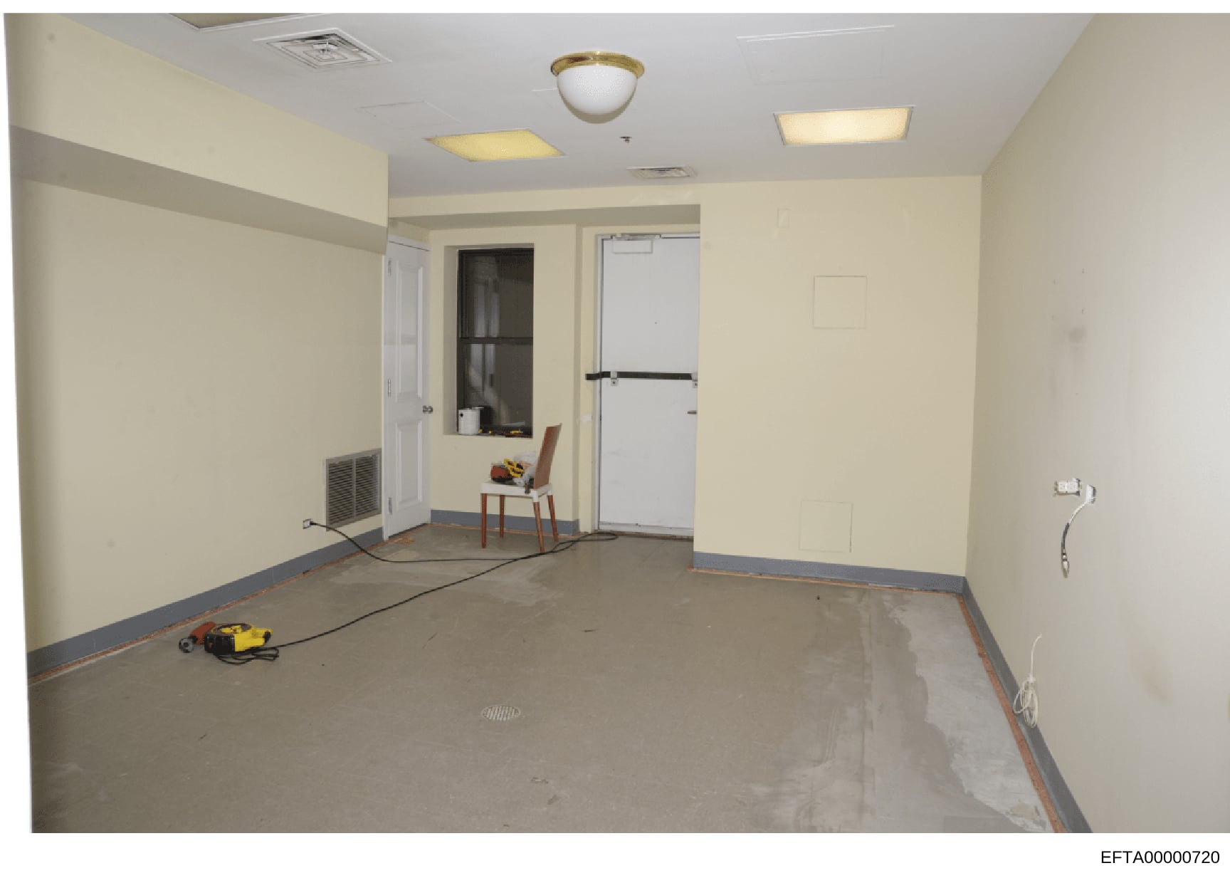 This is an evidence photograph showing an interior room in various stages of renovation or construction. The room appears to be part of a residential or commercial property, with bare concrete flooring, cream-colored walls, recessed lighting, and var