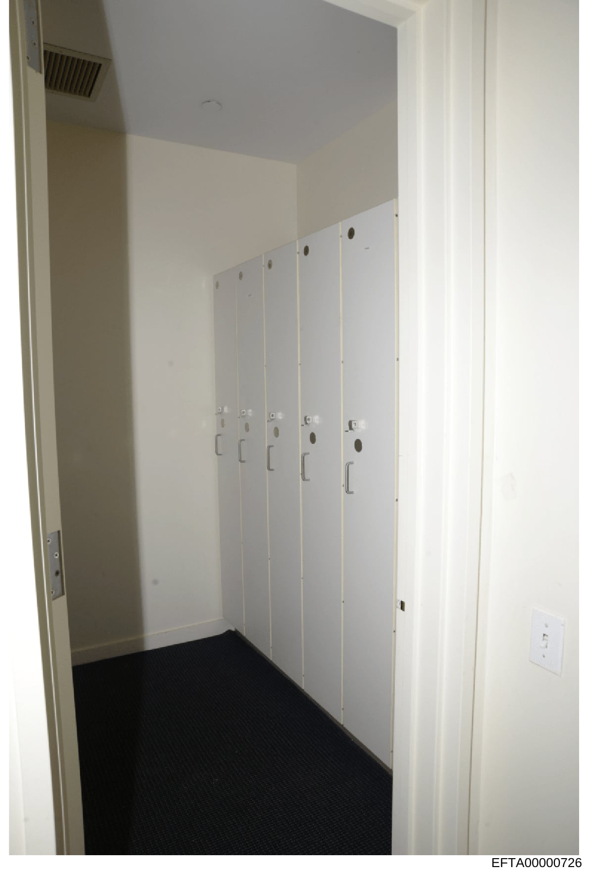 This photograph depicts the interior of a closet or storage room featuring five white locker-style cabinets with metal handles, mounted on walls with beige and blue paint. The room has black carpeting and basic electrical outlets, suggesting this is