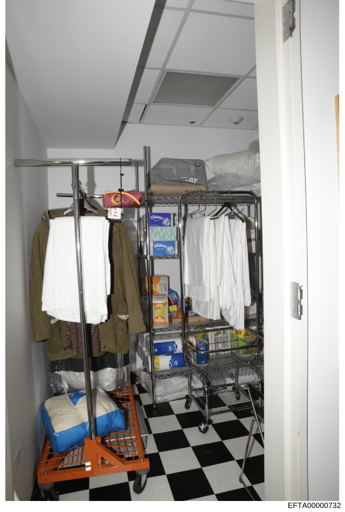 This is a photograph of an interior storage space containing household supplies, linens, clothing, and various packaged goods. The room features metal shelving units, an orange utility cart, white towels, clothing items, and boxes of household produc