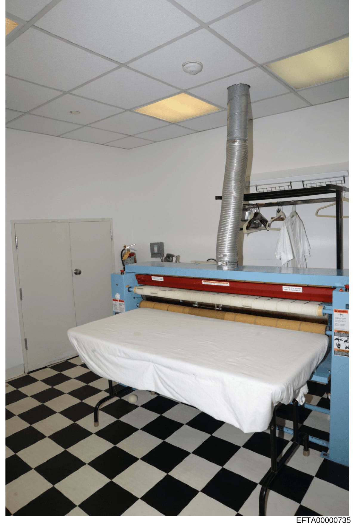 This is a photograph of a laundry or utility room, showing commercial-grade pressing/ironing equipment with a blue frame, white surface covering on a table, and a large metal ductwork pipe extending from the ceiling. The room has a checkered black an