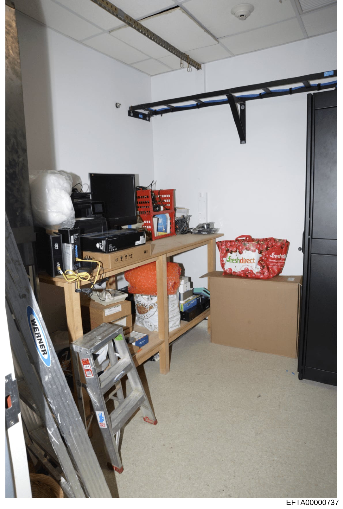 This is an evidence photograph showing an interior room containing a computer workstation, shelving units, and various storage boxes and supplies. The room appears to be a small office or utility space with minimal furnishings. The photograph documen