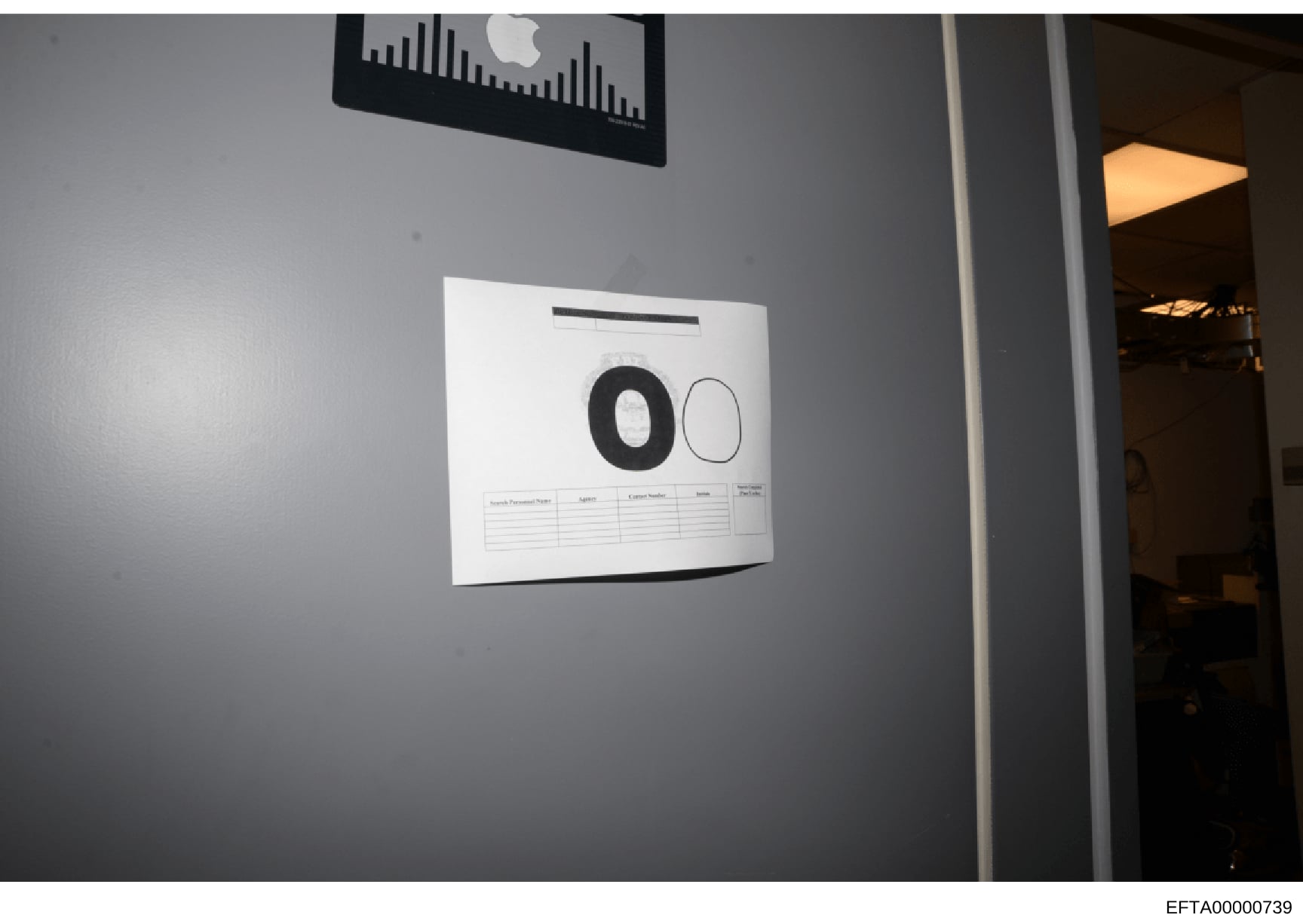 This is an evidence photograph showing a document posted on a gray wall, photographed with scale reference markers (numbered circles '0' and '00'). The document appears to be a form or record with a table below the scale markers. An Apple device is v