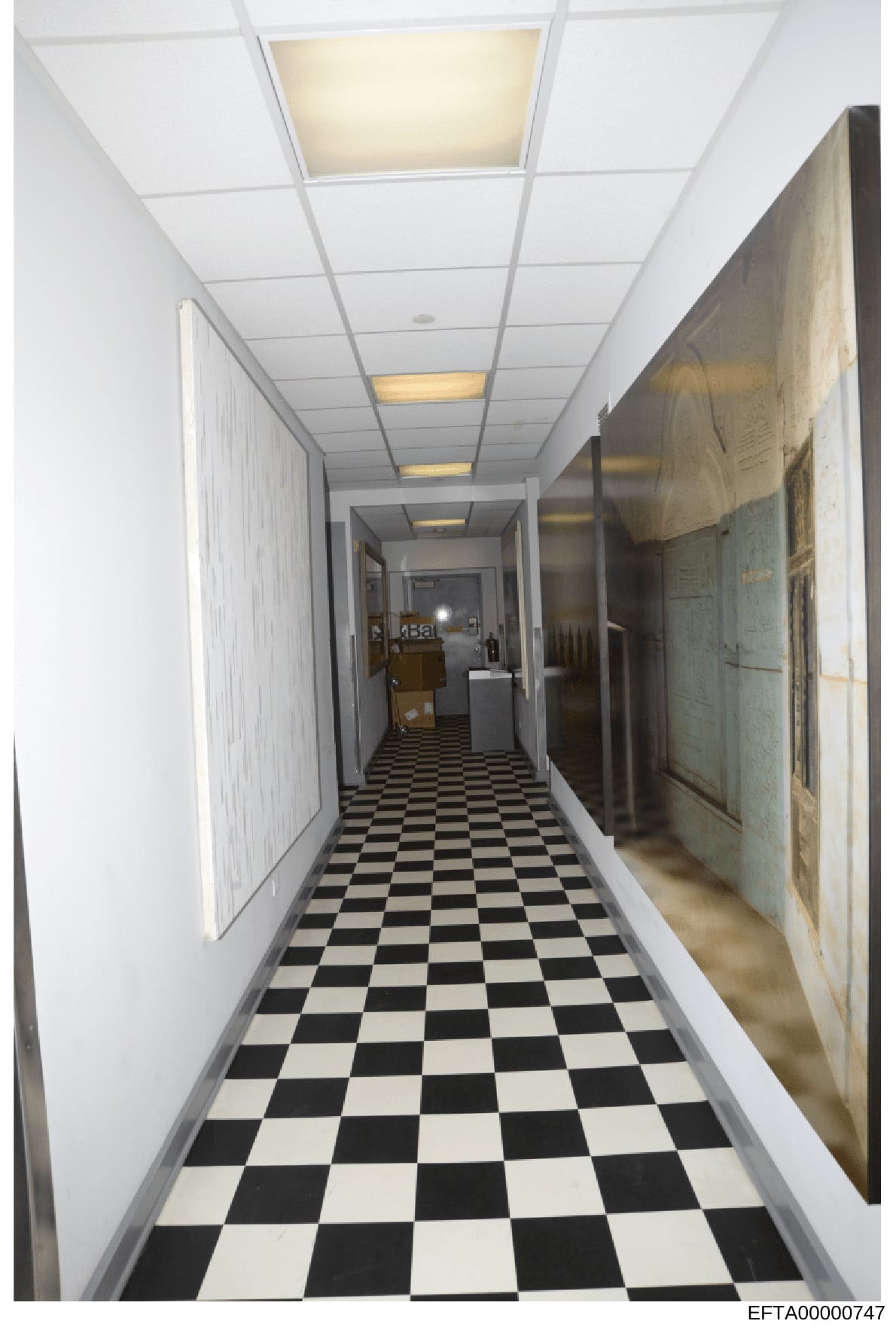 This is a photograph of a narrow, institutional-style hallway featuring a black and white checkered tile floor, white walls, drop-ceiling with recessed lighting, and what appears to be storage or utility items along the right side. The hallway appear