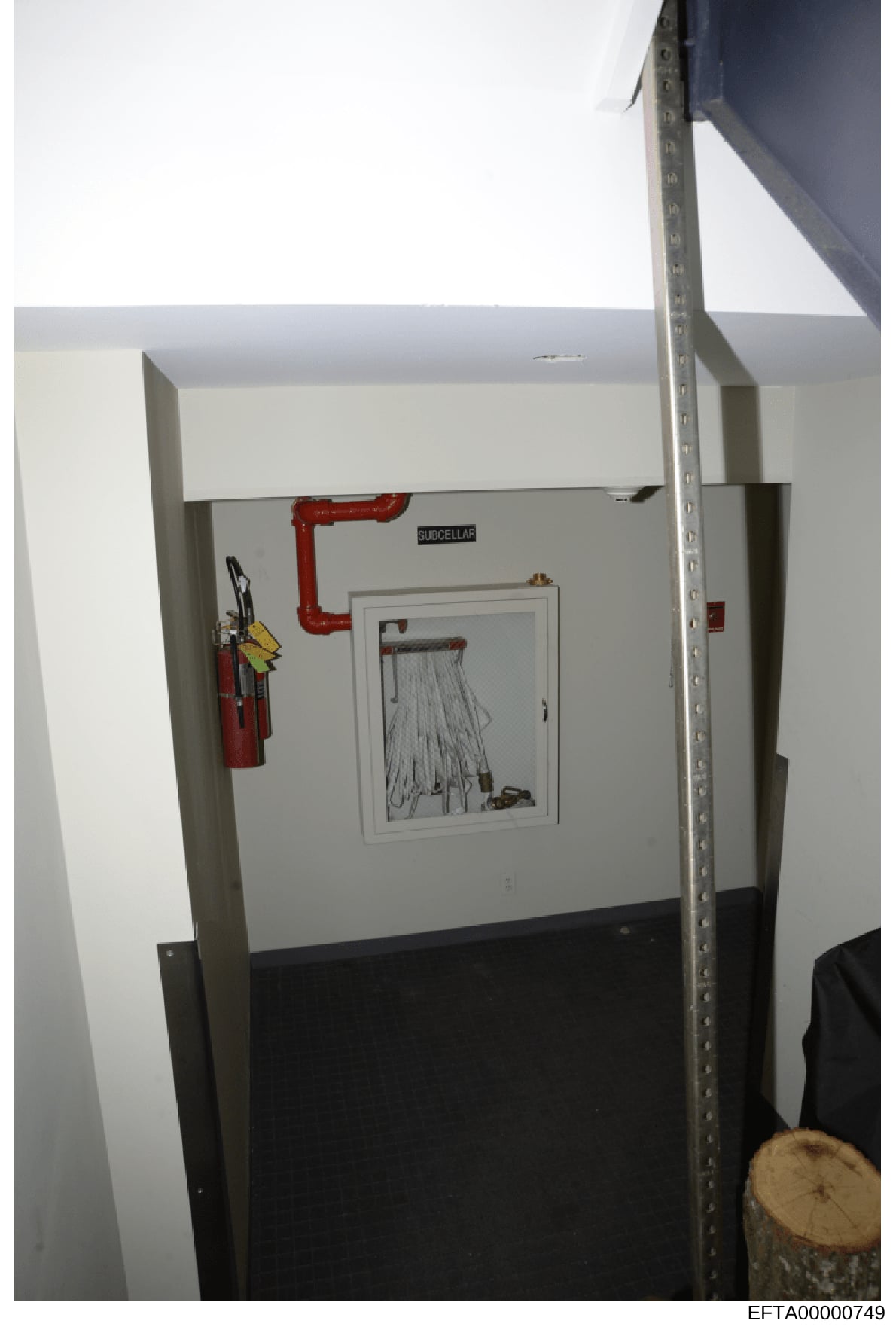 Photograph of an interior utility or mechanical room showing fire suppression and safety equipment. The image depicts a wall-mounted fire extinguisher, red piping labeled 'SUBCELLAR', and an open cabinet containing coiled hoses and maintenance equipm