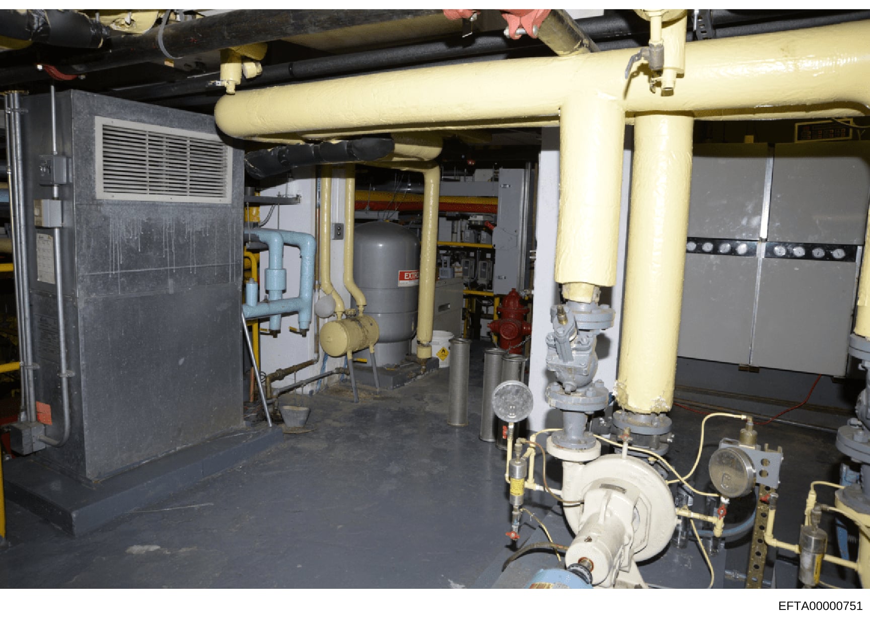 This is a photograph of a basement mechanical room, likely from an Epstein property, showing industrial heating, ventilation, and air conditioning (HVAC) equipment, piping systems, and utility infrastructure. The image documents the building's mechan