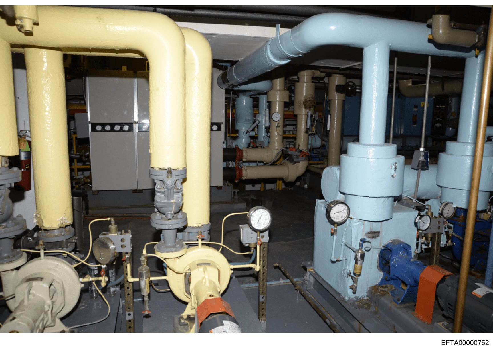 This photograph shows the interior of a mechanical equipment room containing heating, ventilation, and air conditioning (HVAC) systems. The image displays large yellow and light blue piping systems, pressure gauges, control panels, and various mechan