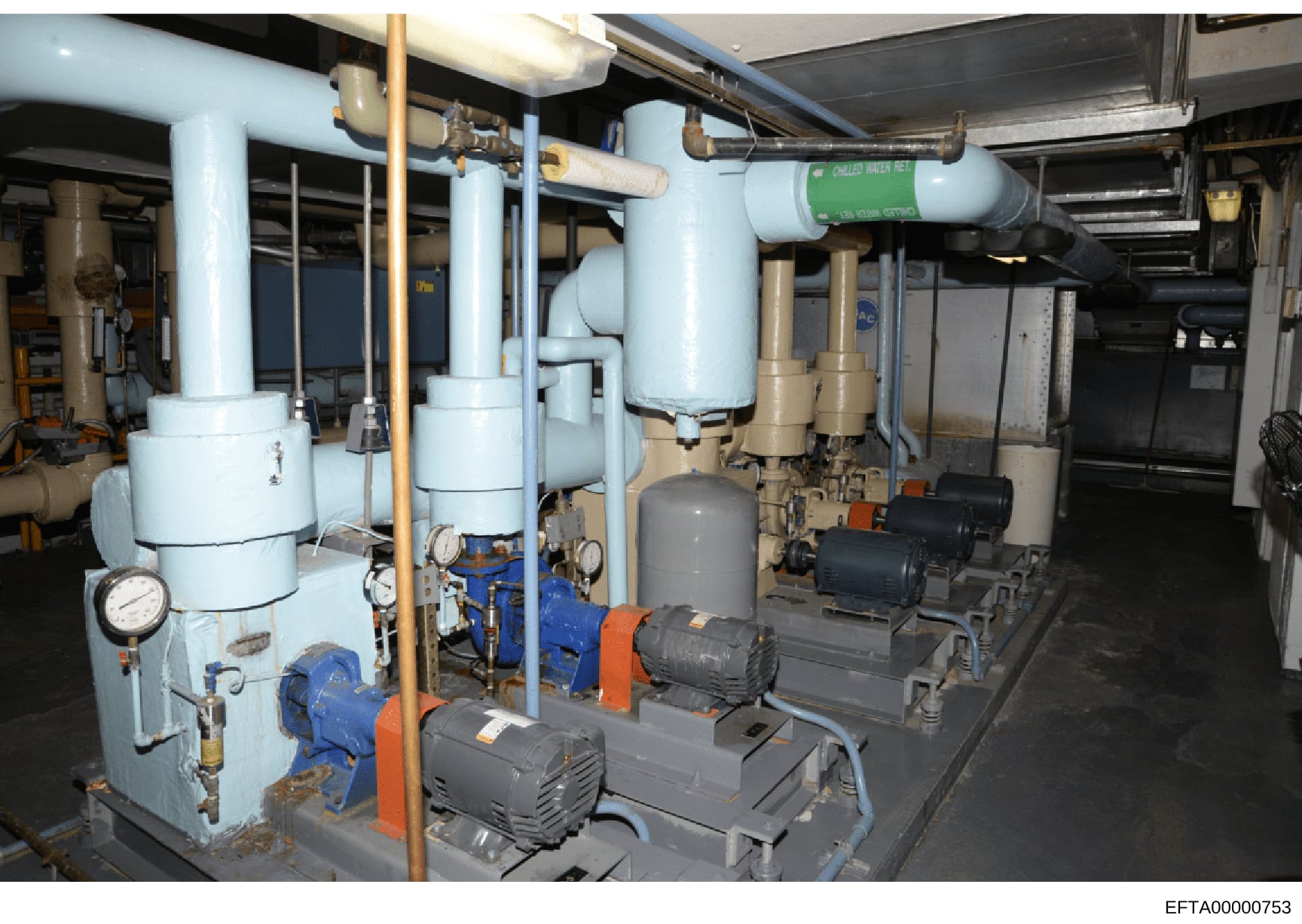 This photograph documents a building's mechanical room containing HVAC (heating, ventilation, and air conditioning) and plumbing infrastructure. The image shows industrial-grade equipment including white cylindrical tanks, blue and black pumps with o