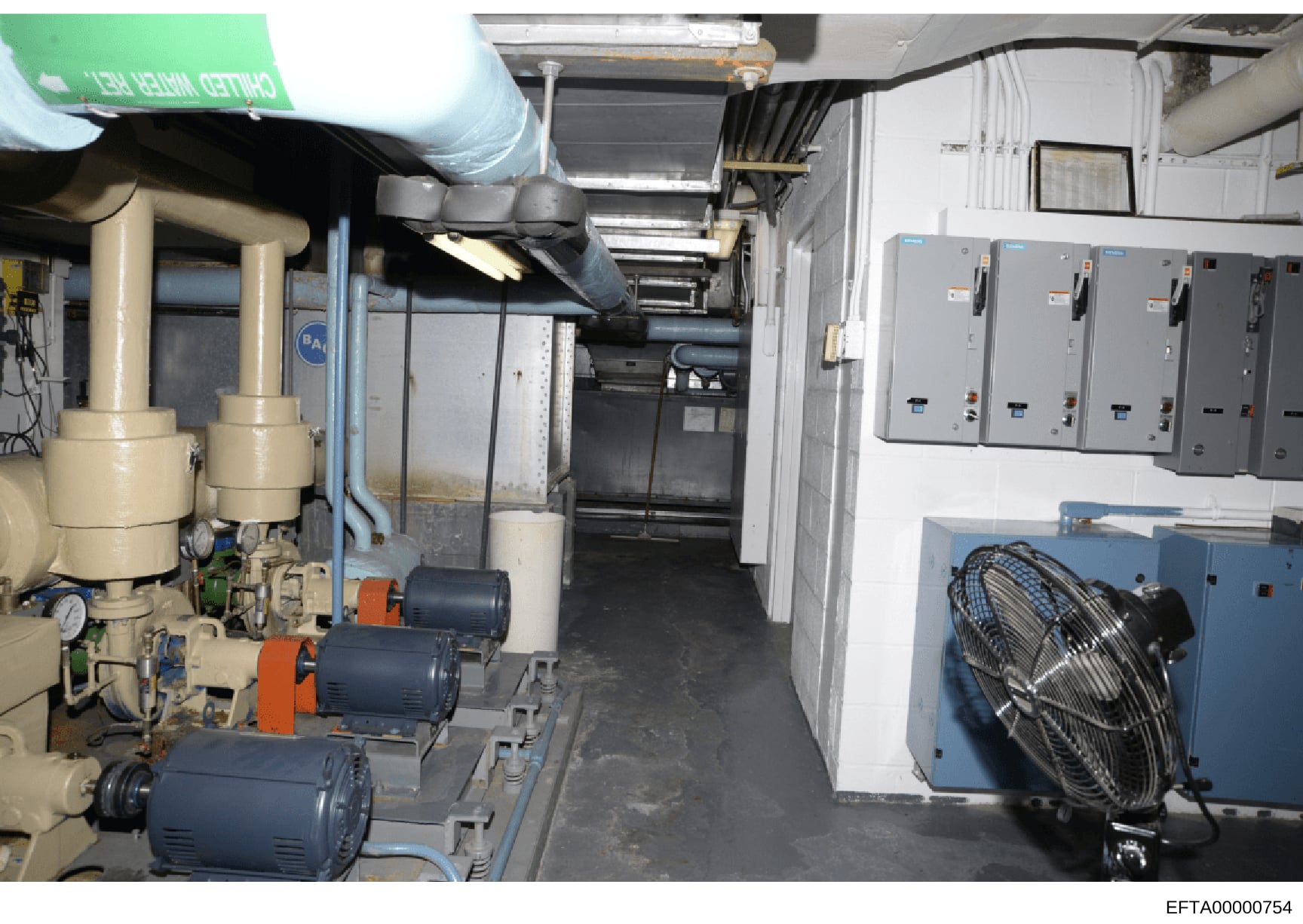 This photograph documents an interior mechanical/utility room containing HVAC equipment, including large tan-colored compressors or condenser units on the left side, multiple electrical control panels mounted on the right wall, ductwork, piping infra