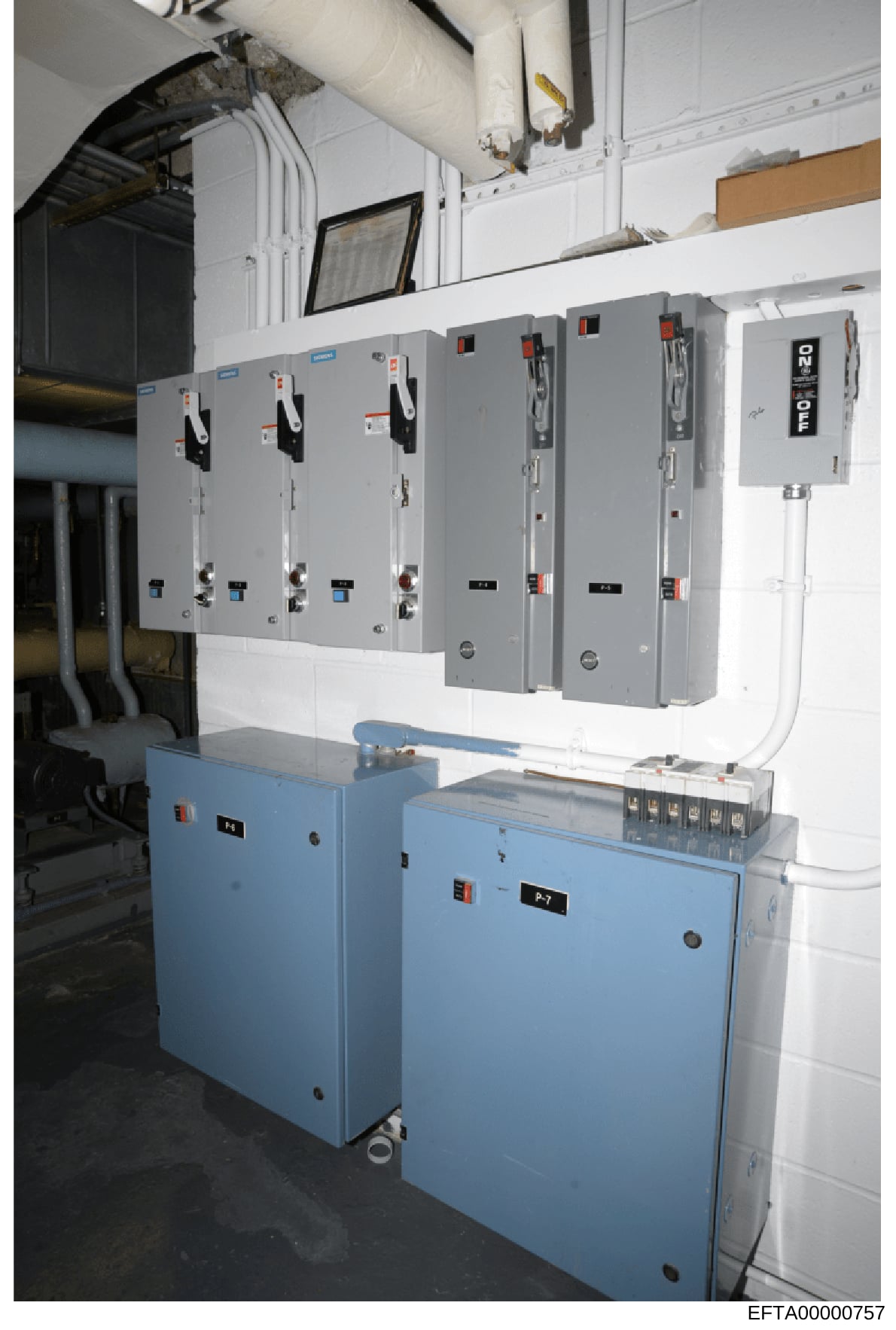 This photograph shows an electrical utility room with multiple gray electrical panels mounted on a white wall above two blue electrical control boxes. The image appears to be from a property inspection or evidence documentation, capturing the electri