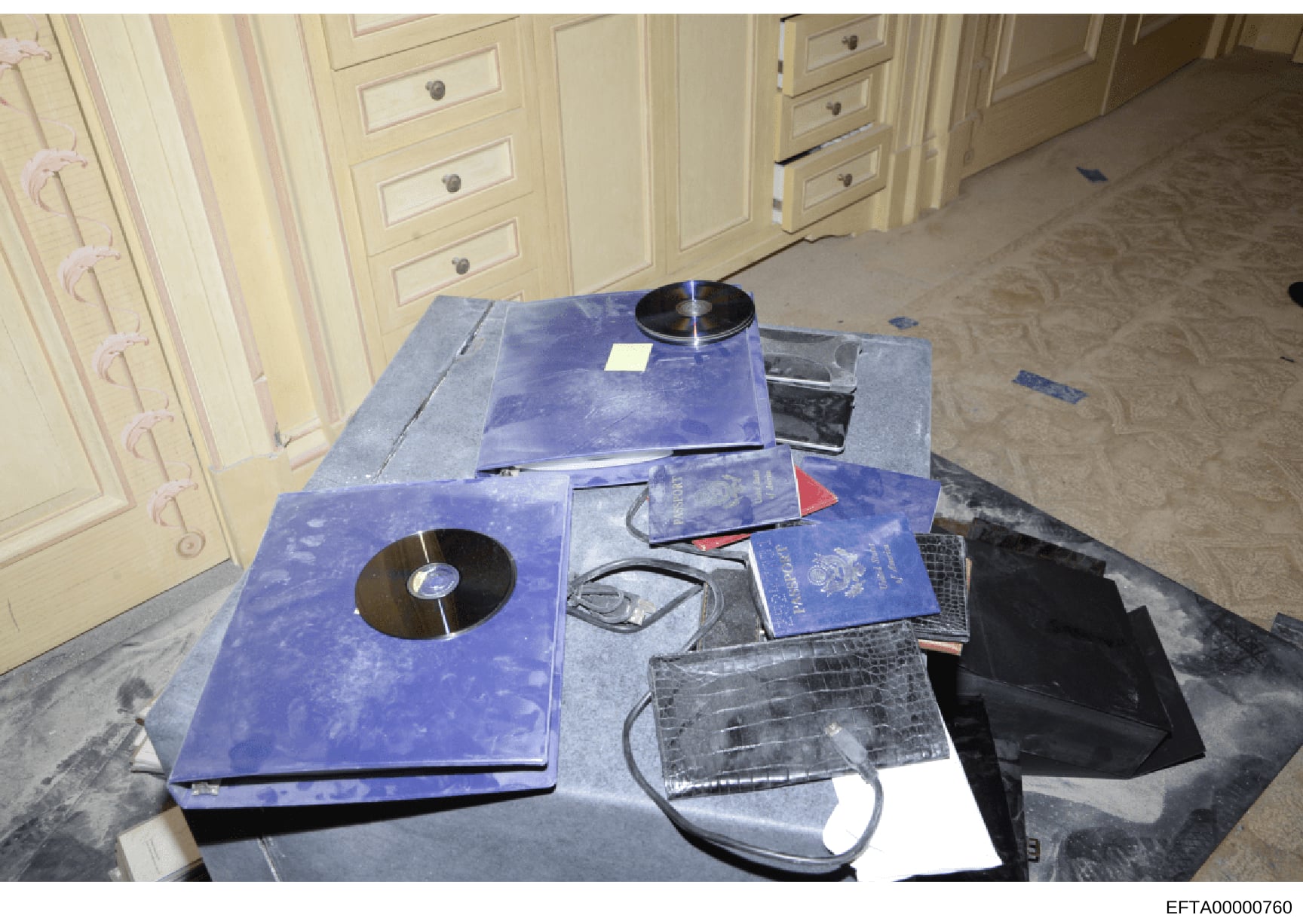 This is an evidence photograph showing items seized during a search, displayed on a table in what appears to be a residential interior. The image shows multiple blue passports, CDs/DVDs, electronic devices, and what appears to be crocodile or alligat