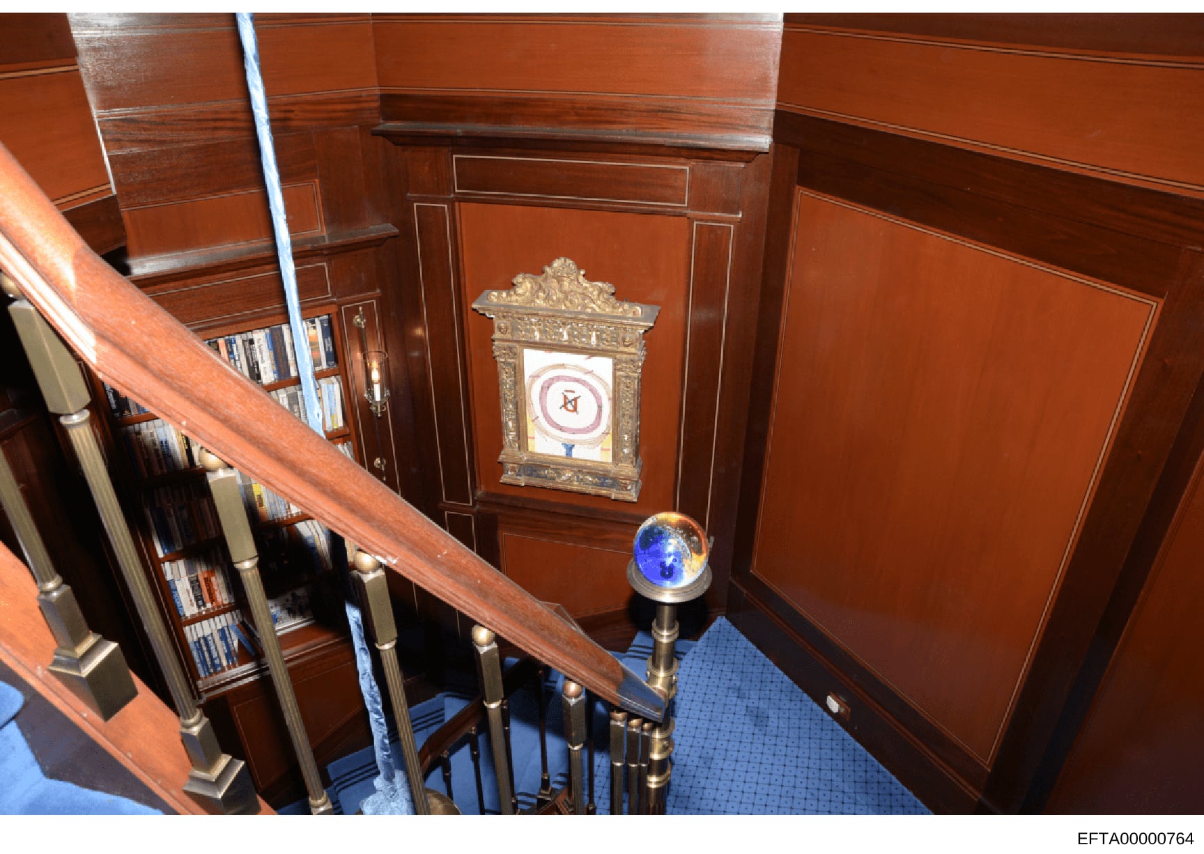 This is an evidence photograph showing the interior of a residential property featuring a wooden staircase with ornate railings, built-in bookshelves, and decorative items including a framed heraldic emblem and a blue globe. The photo documents the a