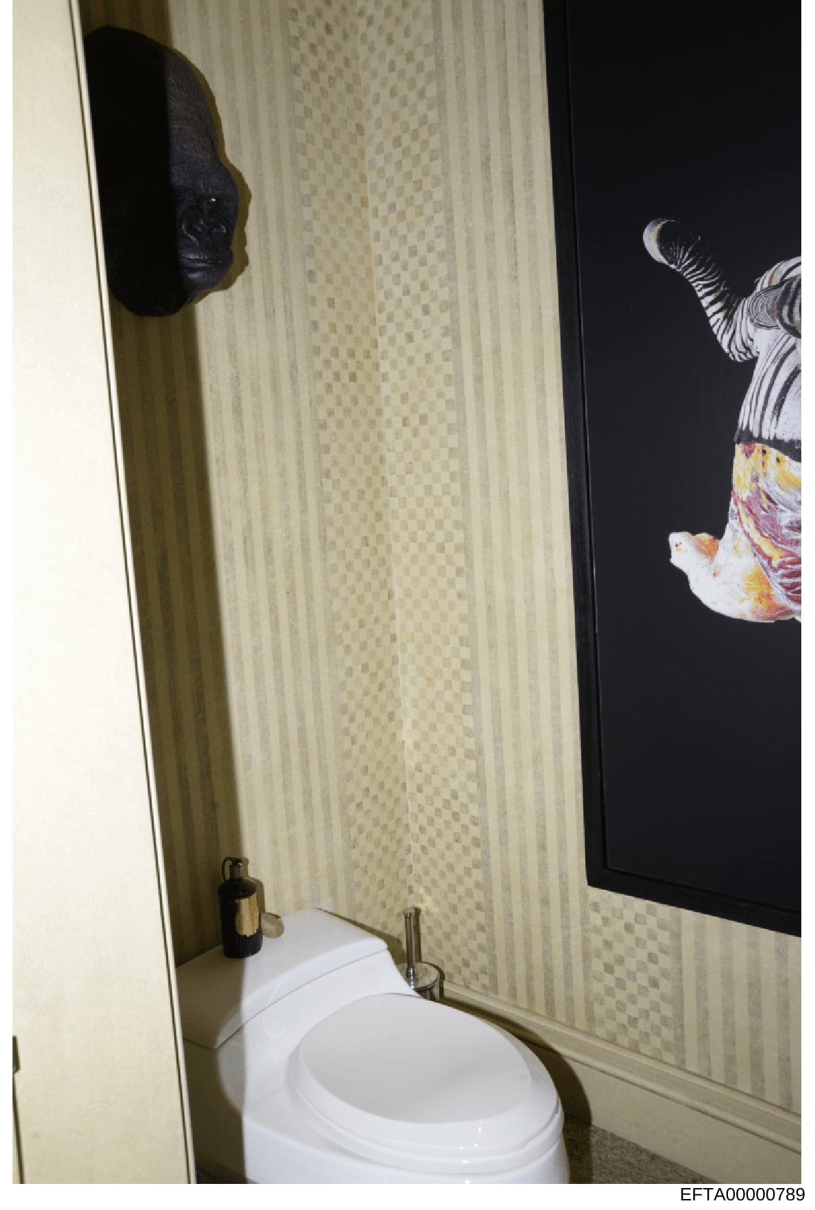 This photograph shows the interior of a bathroom, likely from one of Jeffrey Epstein's properties. The image captures a white toilet, cream-colored tiled walls with decorative striping, framed artwork including a black sculptural mask and a colorful 
