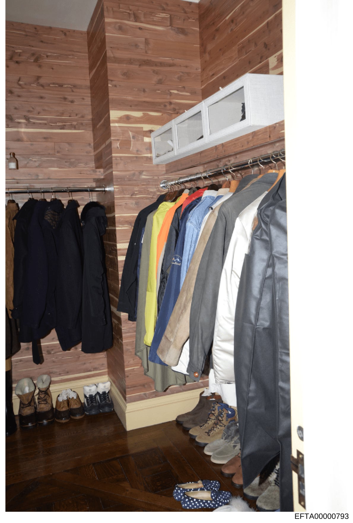 This is an evidence photograph showing the interior of a walk-in closet with wooden paneled walls. The closet contains multiple clothing items on hanging rods (including jackets, coats, and other garments in various colors) and shoes/boots organized 