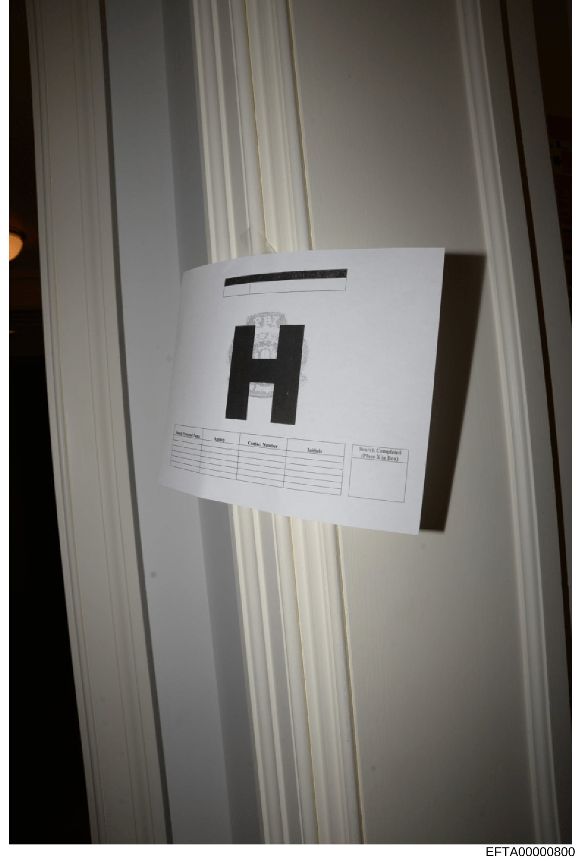 This is a photograph of evidence documentation showing a large black letter 'H' with an evidence label attached, positioned against white curtains or drapes. The label appears to be part of a formal evidence collection and cataloging procedure, with 