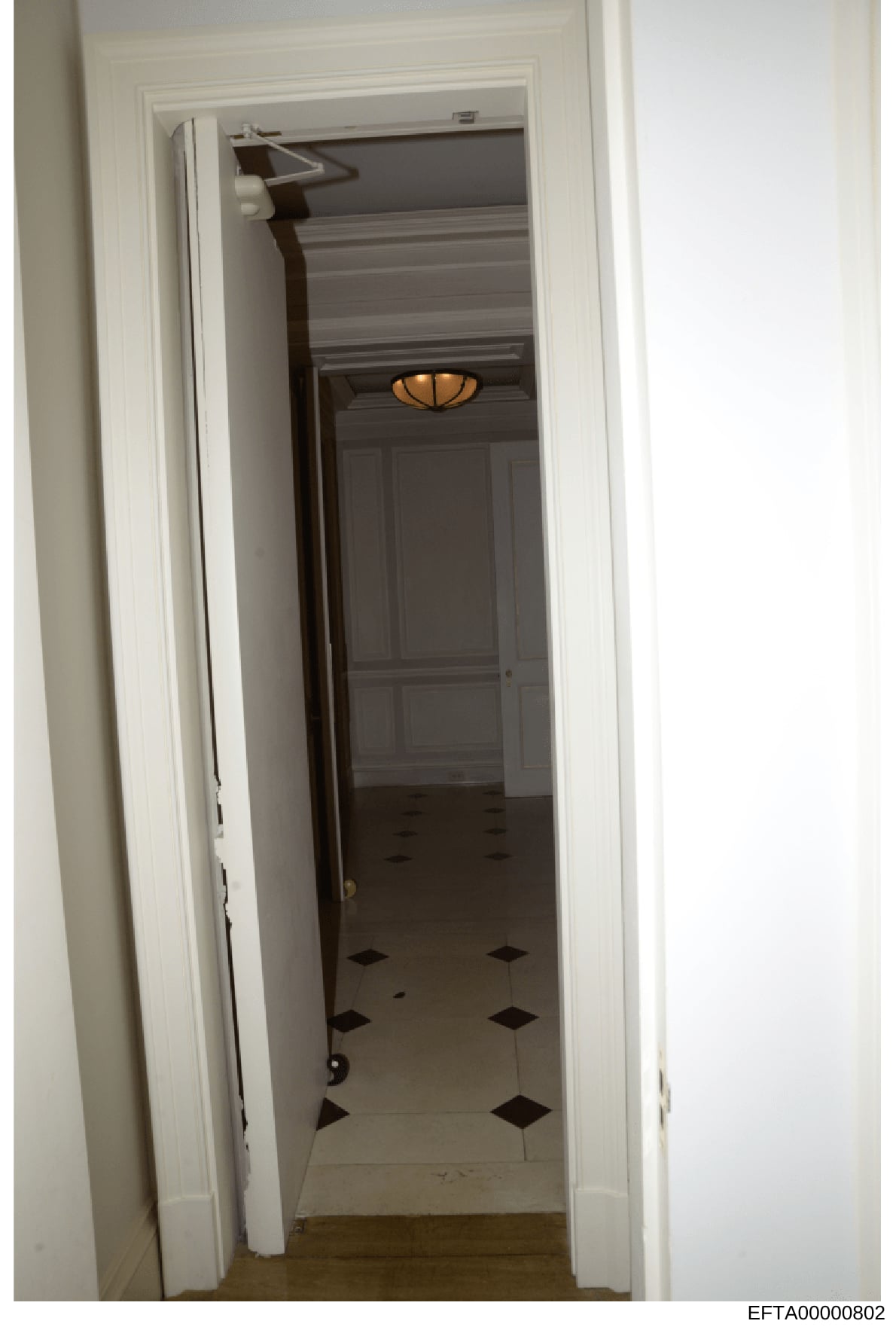 This is a photograph of an interior hallway, likely from one of Jeffrey Epstein's properties, showing a narrow corridor with white-painted walls and a distinctive black-and-white diamond-patterned floor. A dark wooden door is visible at the end of th