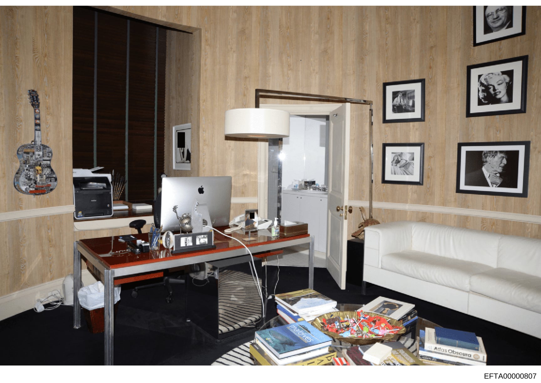 This is a photograph of an upscale bedroom with integrated office area, featuring contemporary furnishings, artwork, and personal items. The room appears to be from one of Jeffrey Epstein's properties and serves as evidence documentation of the inter