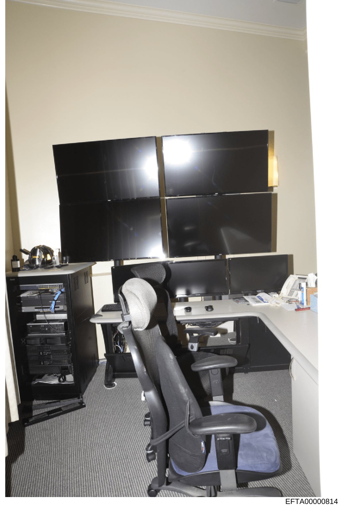 This is a photograph of a professional office workstation featuring multiple computer monitors, server equipment, and office furniture. The image shows a desk setup with six black monitors arranged in a grid pattern, computer server hardware in a rac