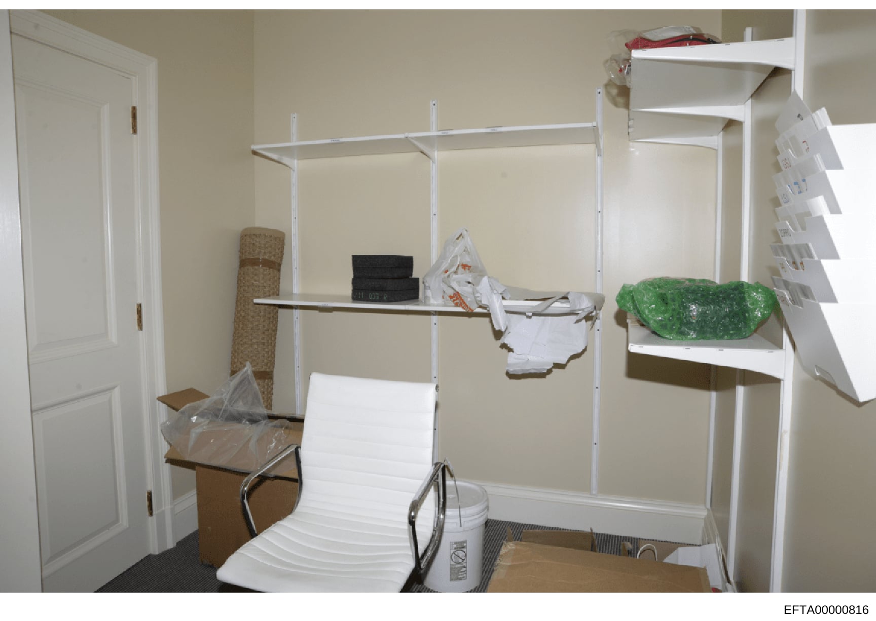 This photograph documents an interior room space containing white metal shelving units with various stored items including boxes, clothing, household goods, and other materials. The image appears to be evidence documentation from a property search or