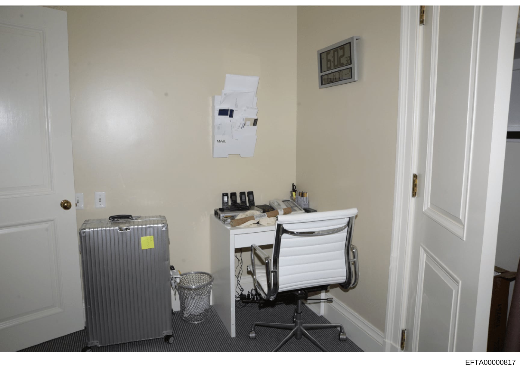 This is an evidence photograph showing an interior office or workspace, likely from one of Epstein's properties. The image captures a small office area with a desk, office chair, filing/mail station on the wall, a wheeled suitcase, waste basket, digi