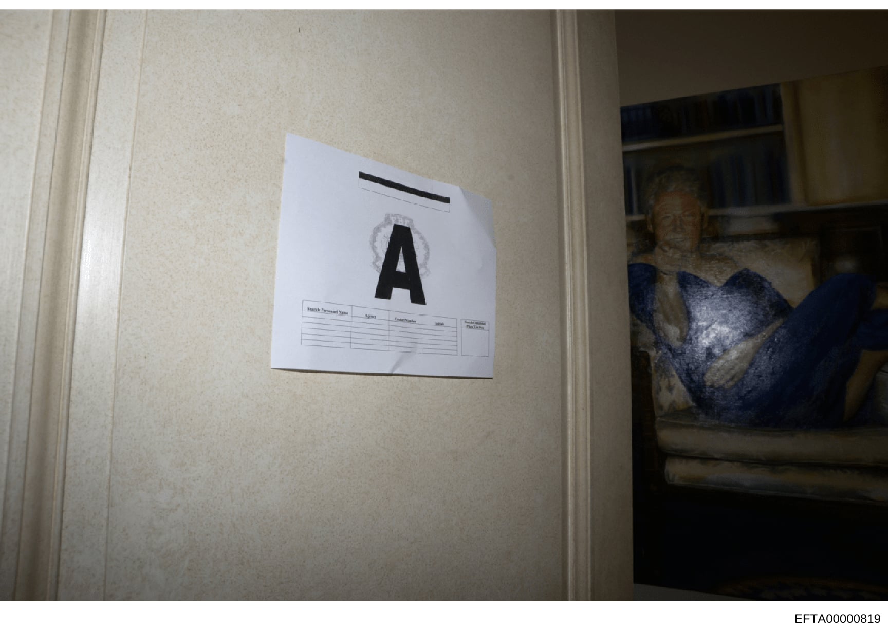 This is an evidence photograph taken during a law enforcement search or inspection, showing a white evidence marker labeled 'A' affixed to a beige/cream-colored wall. The marker includes a documentation grid beneath the letter. A framed photograph is