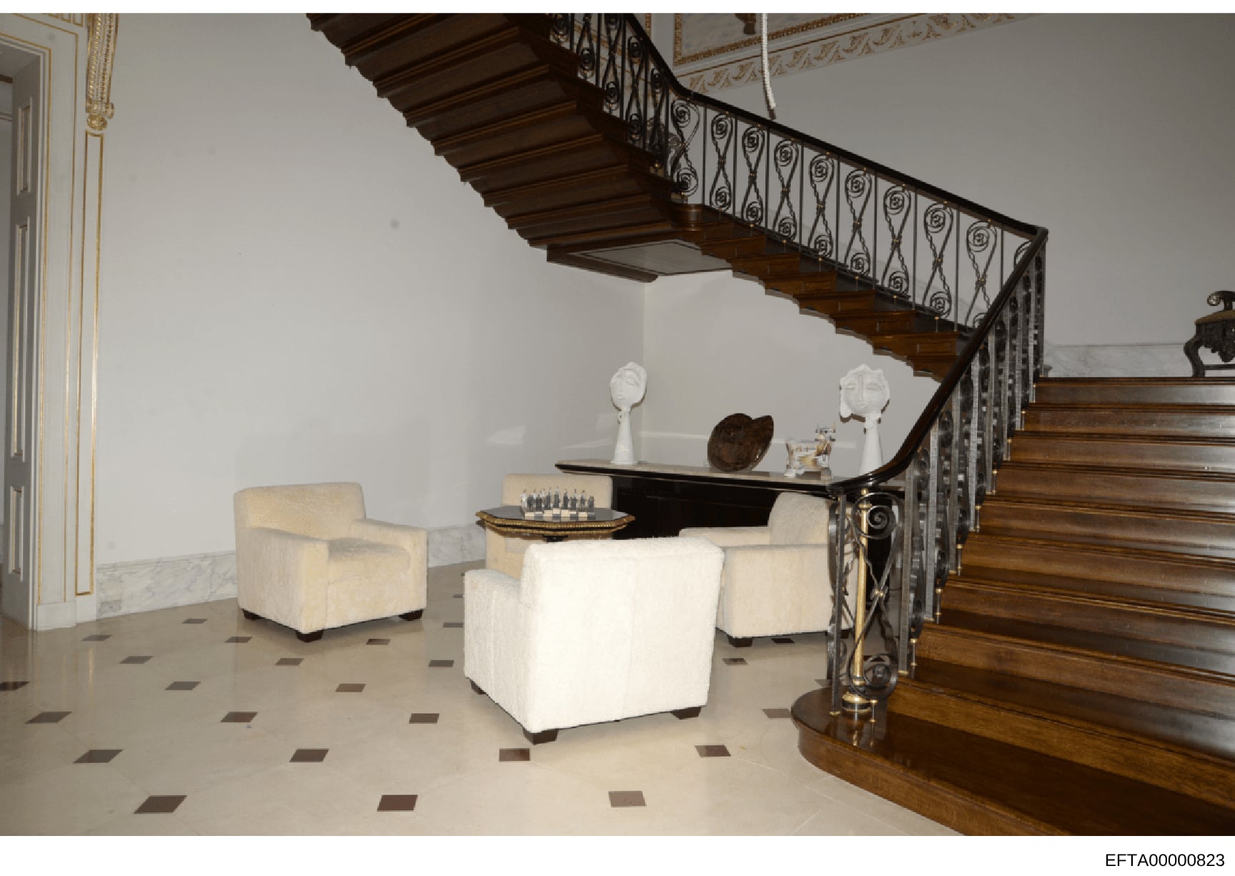 This is an interior photograph of a luxurious residential foyer featuring a curved wooden staircase with ornate iron railing, marble and tile flooring, cream-colored upholstered seating, and decorative sculptures and art objects. The photograph appea
